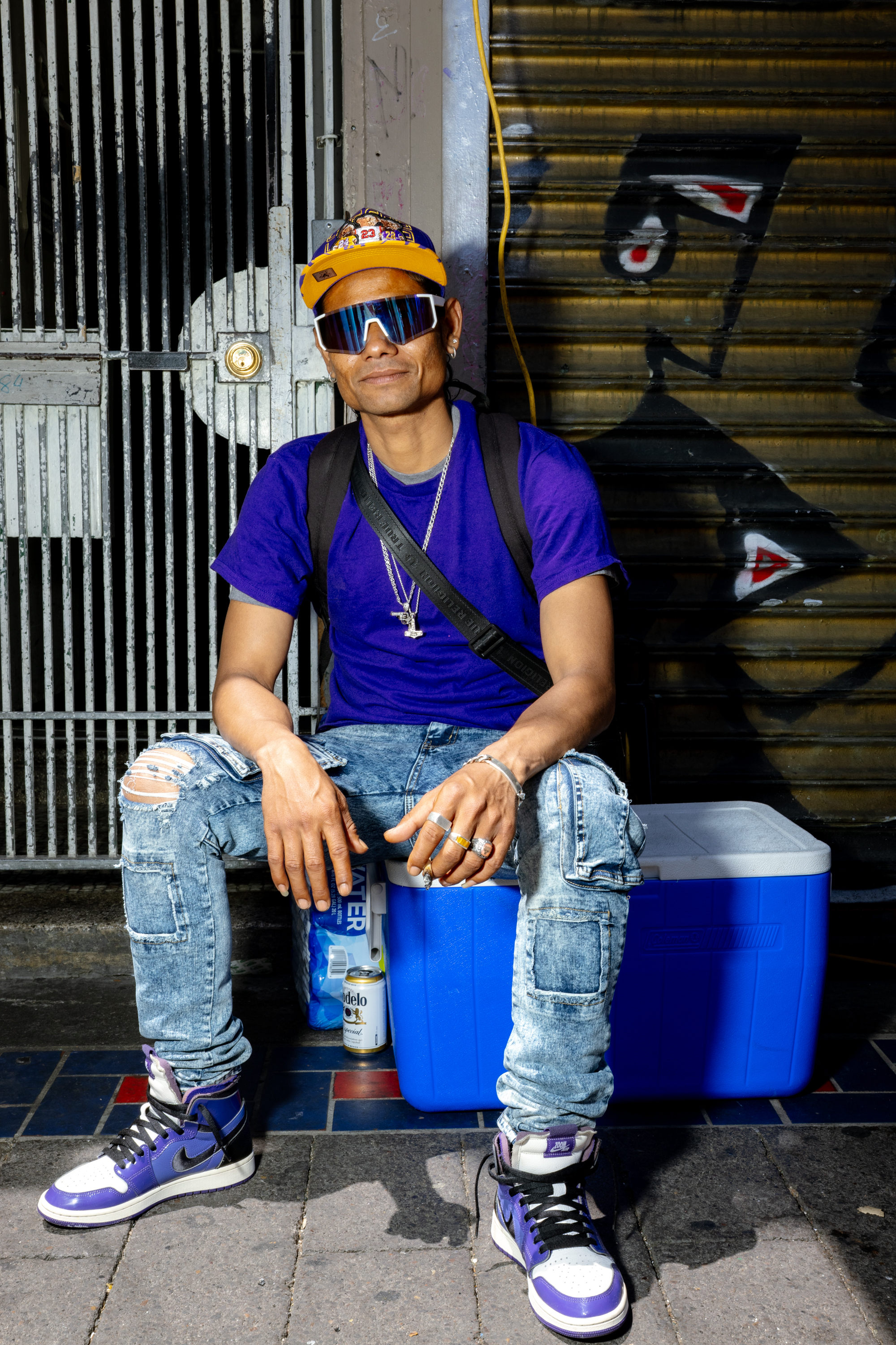 A person in a purple shirt, ripped jeans, and a yellow cap sits on a blue cooler. They wear large sunglasses, sneakers, and various jewelry, with graffiti behind them.