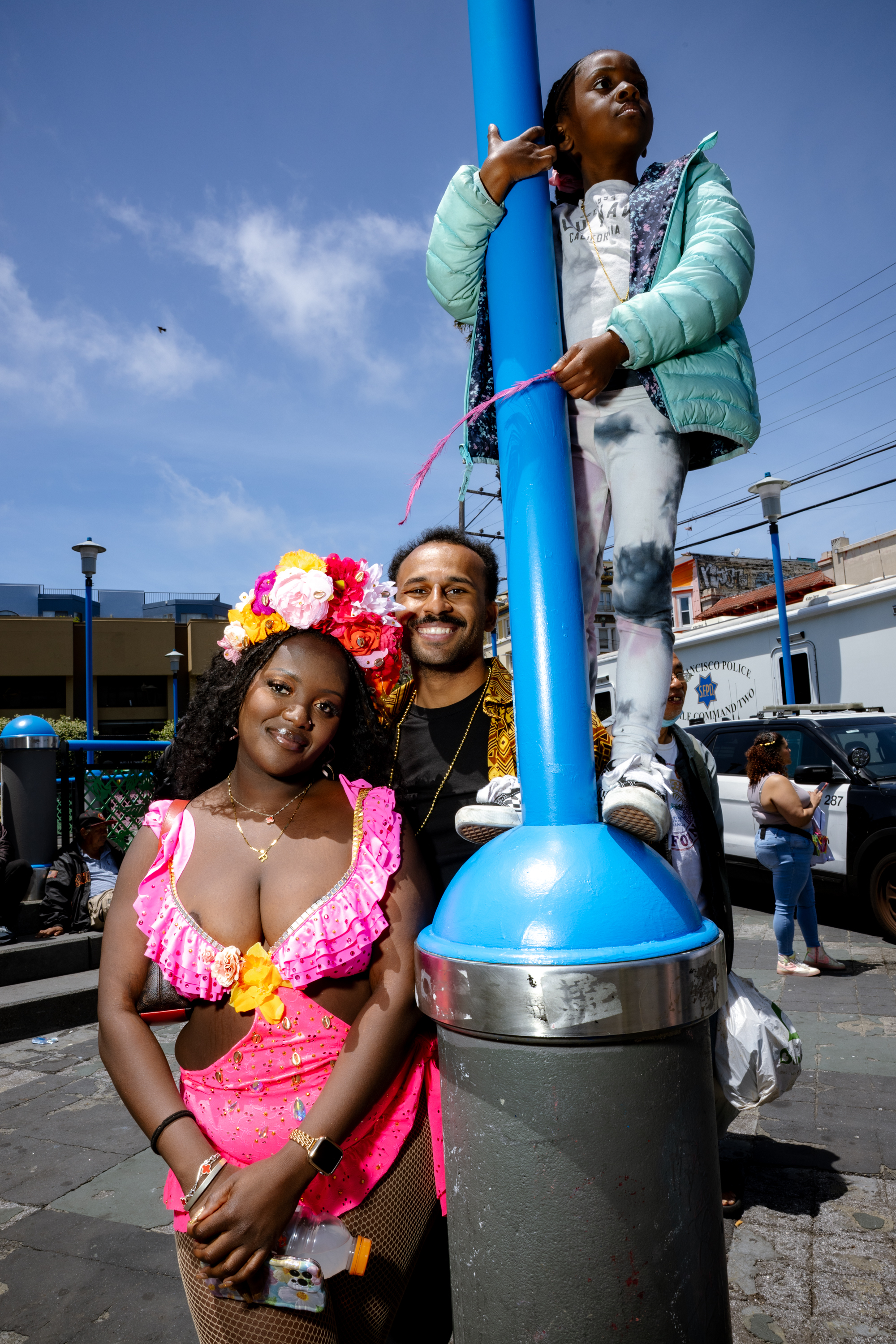 A woman in a vibrant pink outfit smiles with a man and child by a blue pole. The background shows an urban setting under a clear blue sky.