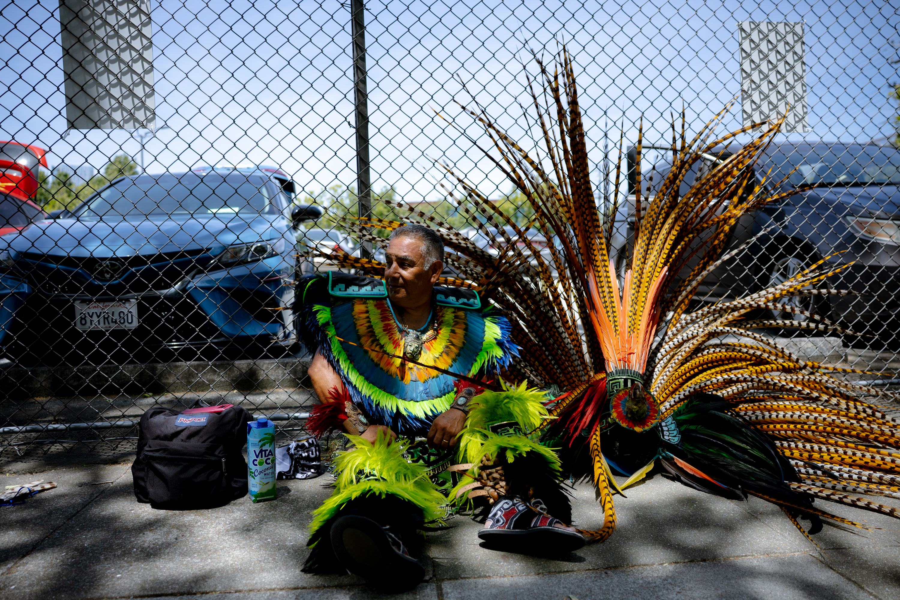 The image shows a man sitting against a chain-link fence, wearing vibrant, colorful traditional attire with feathered decorations. Cars and bags are nearby.