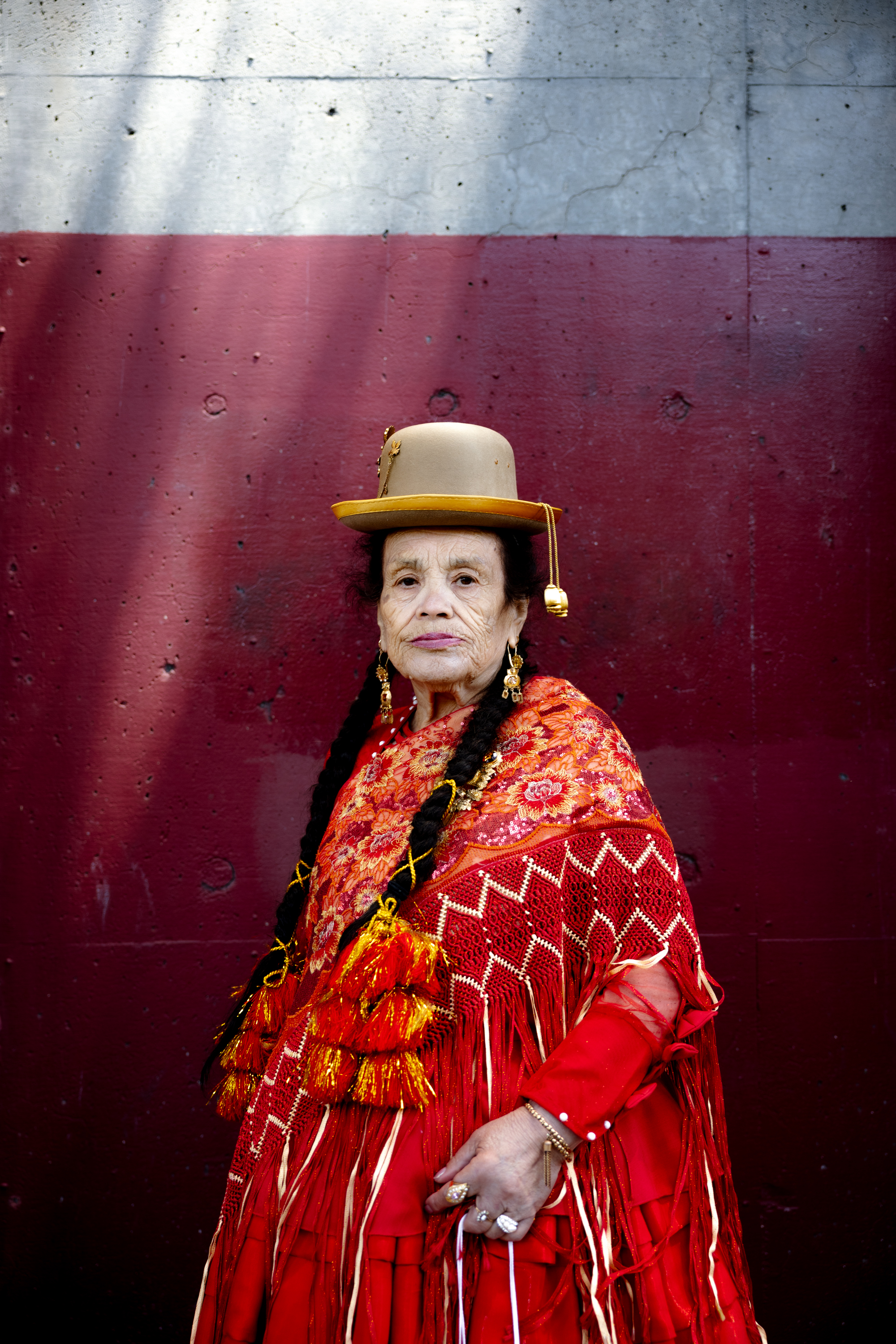 An elderly woman in a red, intricately patterned dress and yellow hat stands against a maroon and gray wall. She has long braids and wears gold earrings.