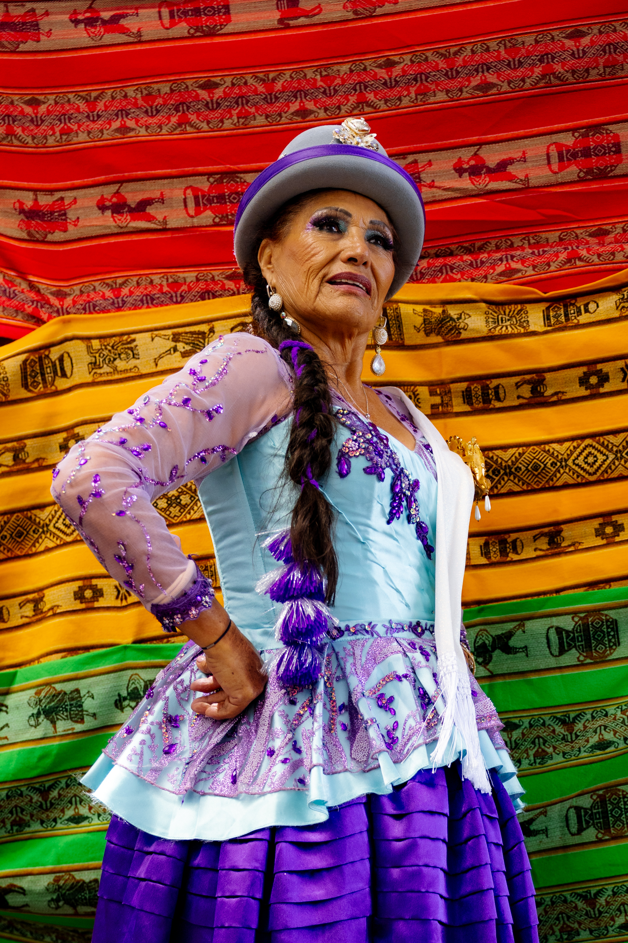 A woman in traditional attire stands confidently, wearing a colorful dress with purple sequins, a hat, and braided hair, against a vibrant, patterned backdrop.