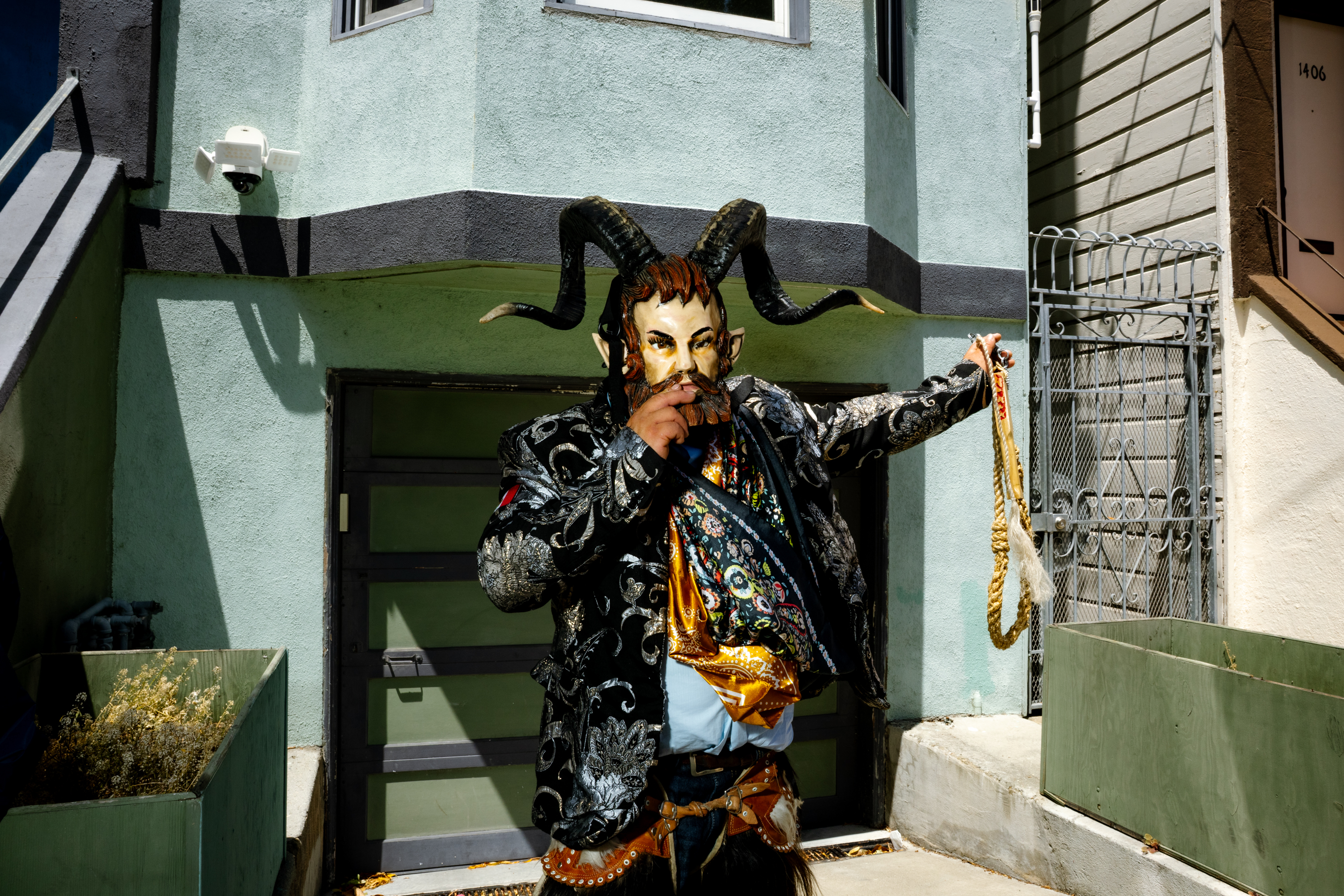 A person stands outside, wearing an ornate, horned mask and a detailed, patterned outfit. They hold a rope, posed in front of a green, urban building.