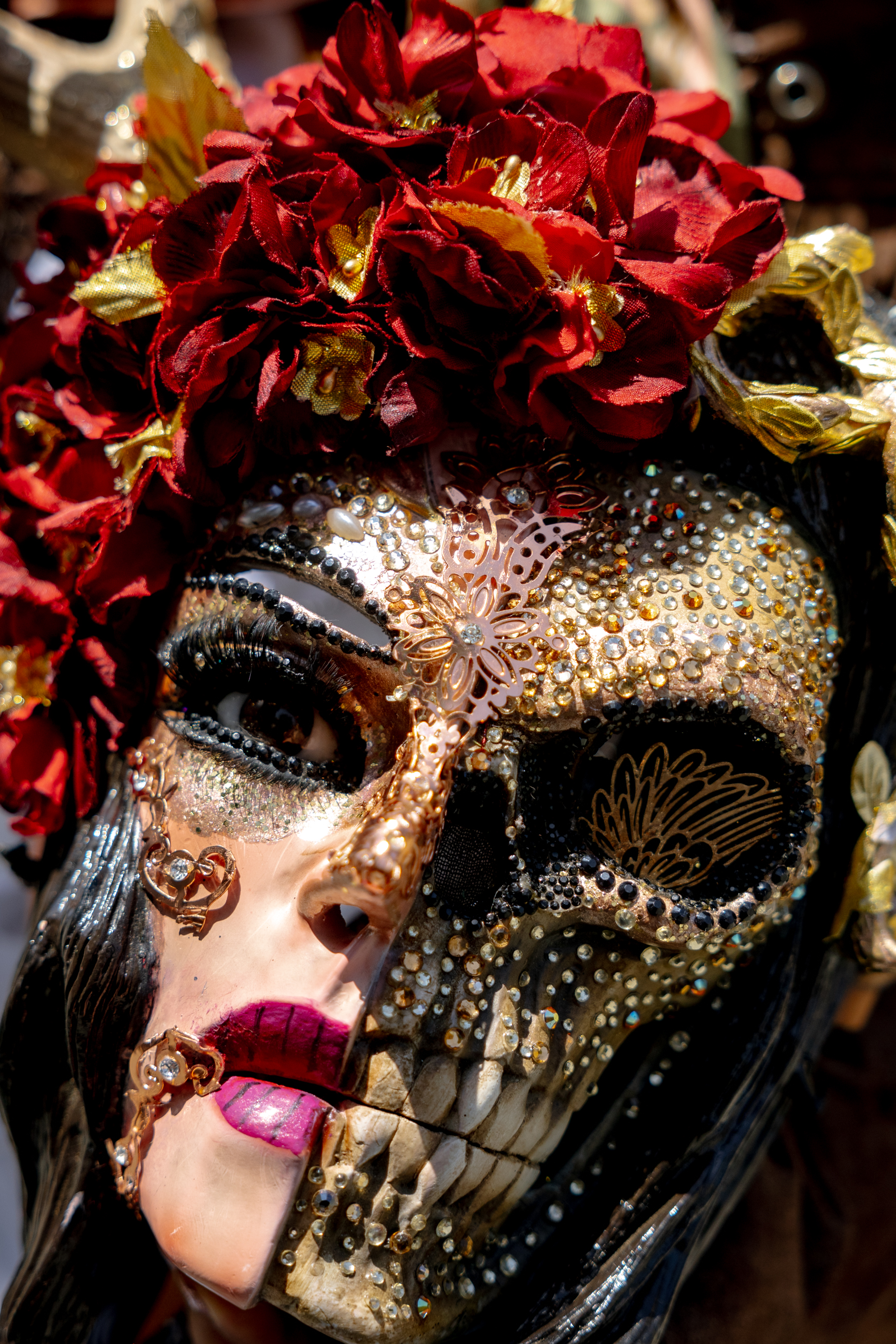 The image shows a lavishly decorated mask, half human, half skeletal, adorned with ornate details, gems, and red flowers, symbolizing a vivid artistic theme.