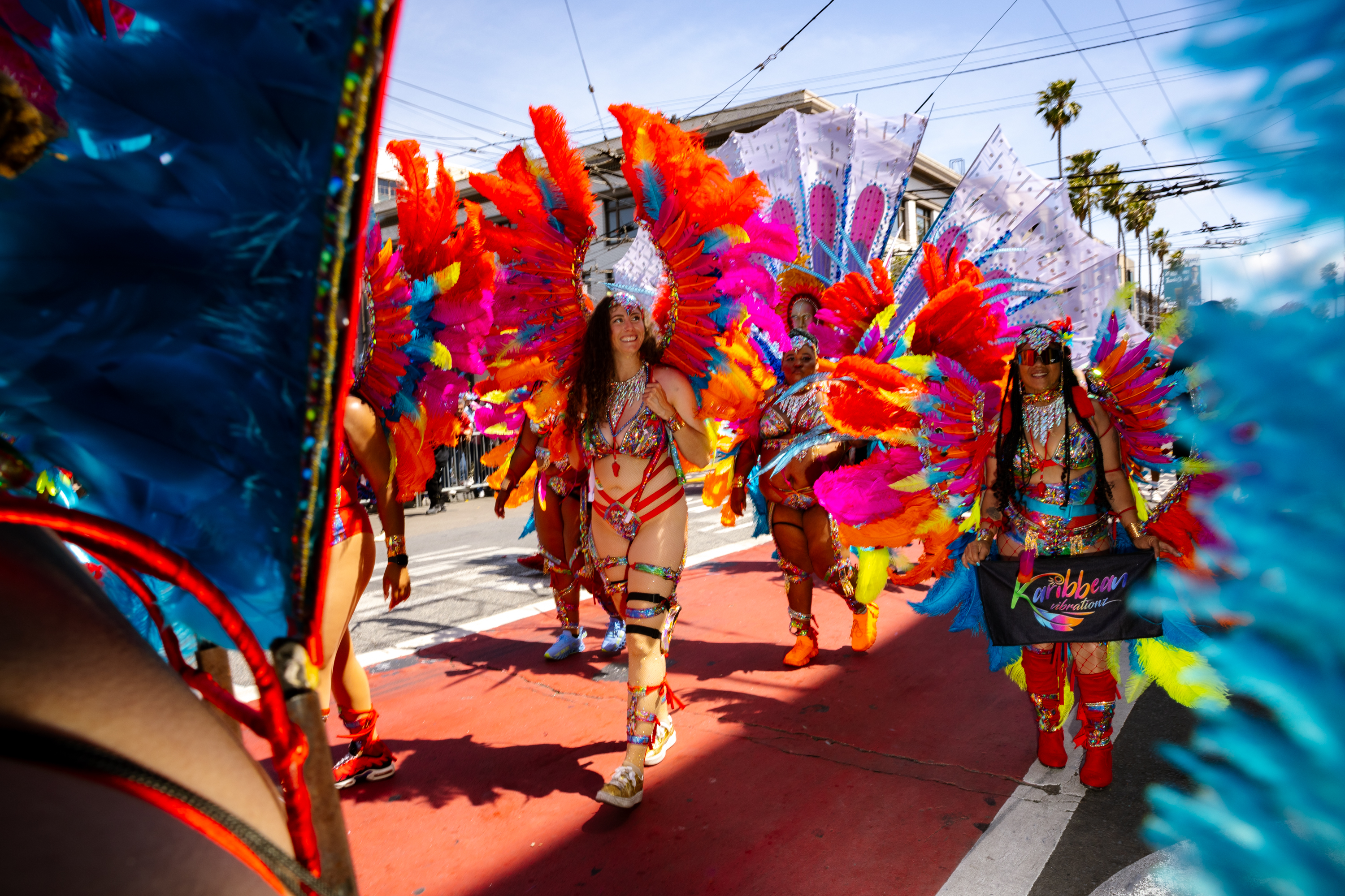 Dancers in vibrant, feathered costumes parade on a street. They wear colorful outfits with large feathered wings, under a sunny blue sky.
