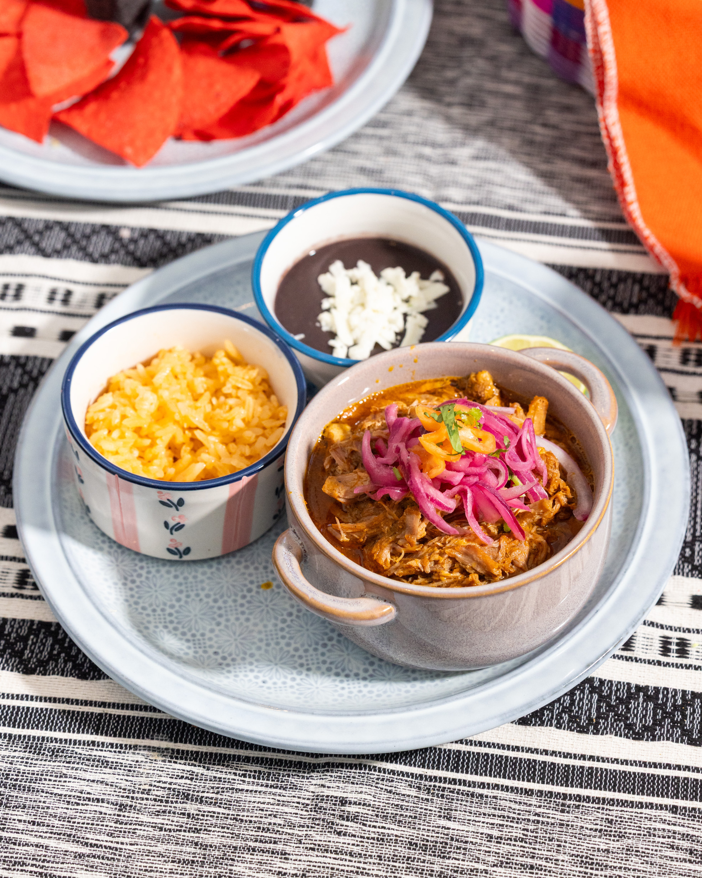 A plate with three bowls: shredded meat with pickled onions, yellow rice, and black beans with cheese. Red tortilla chips are in the background.