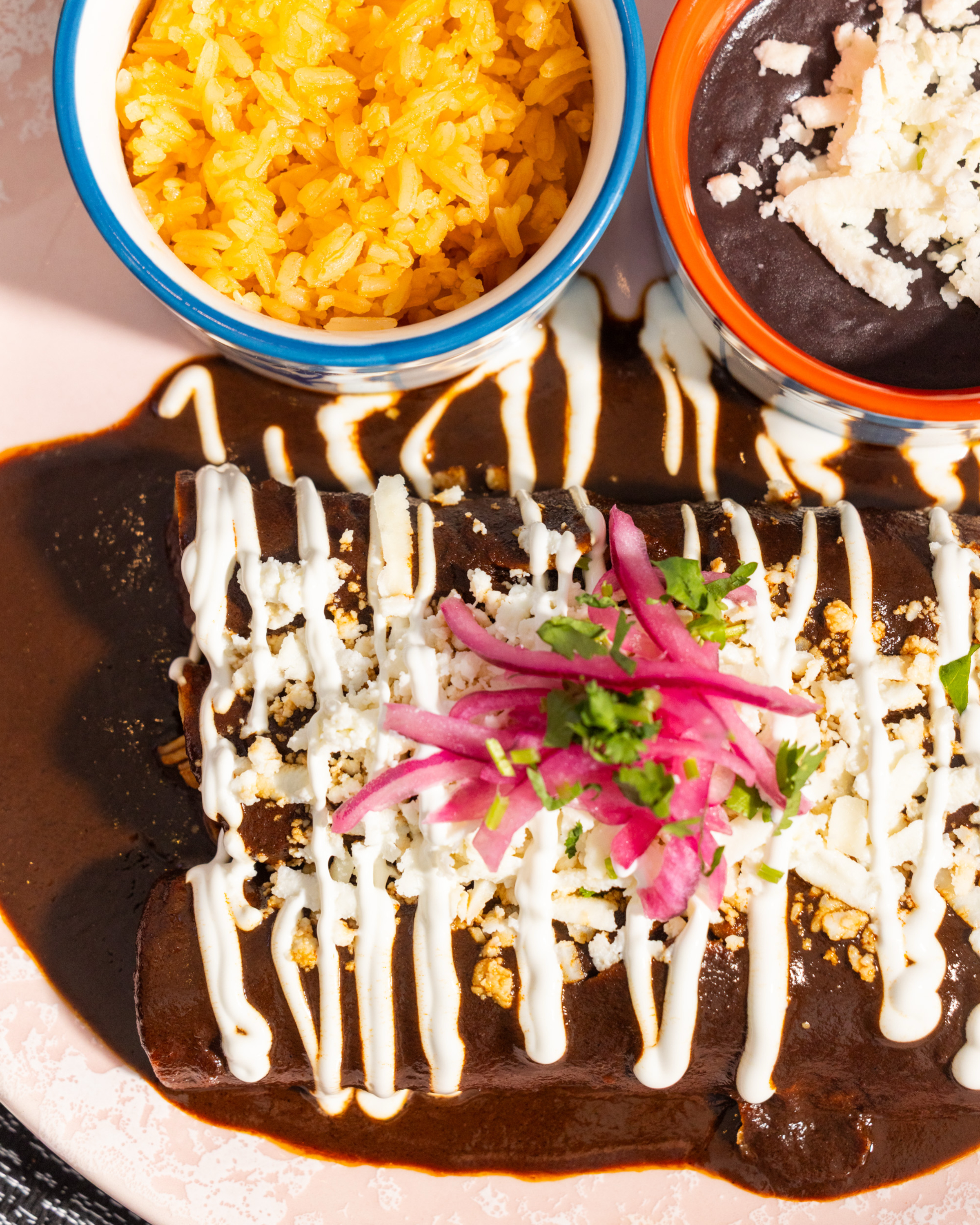 The image shows enchiladas drizzled with cream, topped with cheese and pink pickled onions, alongside bowls of yellow rice and black beans with cheese.