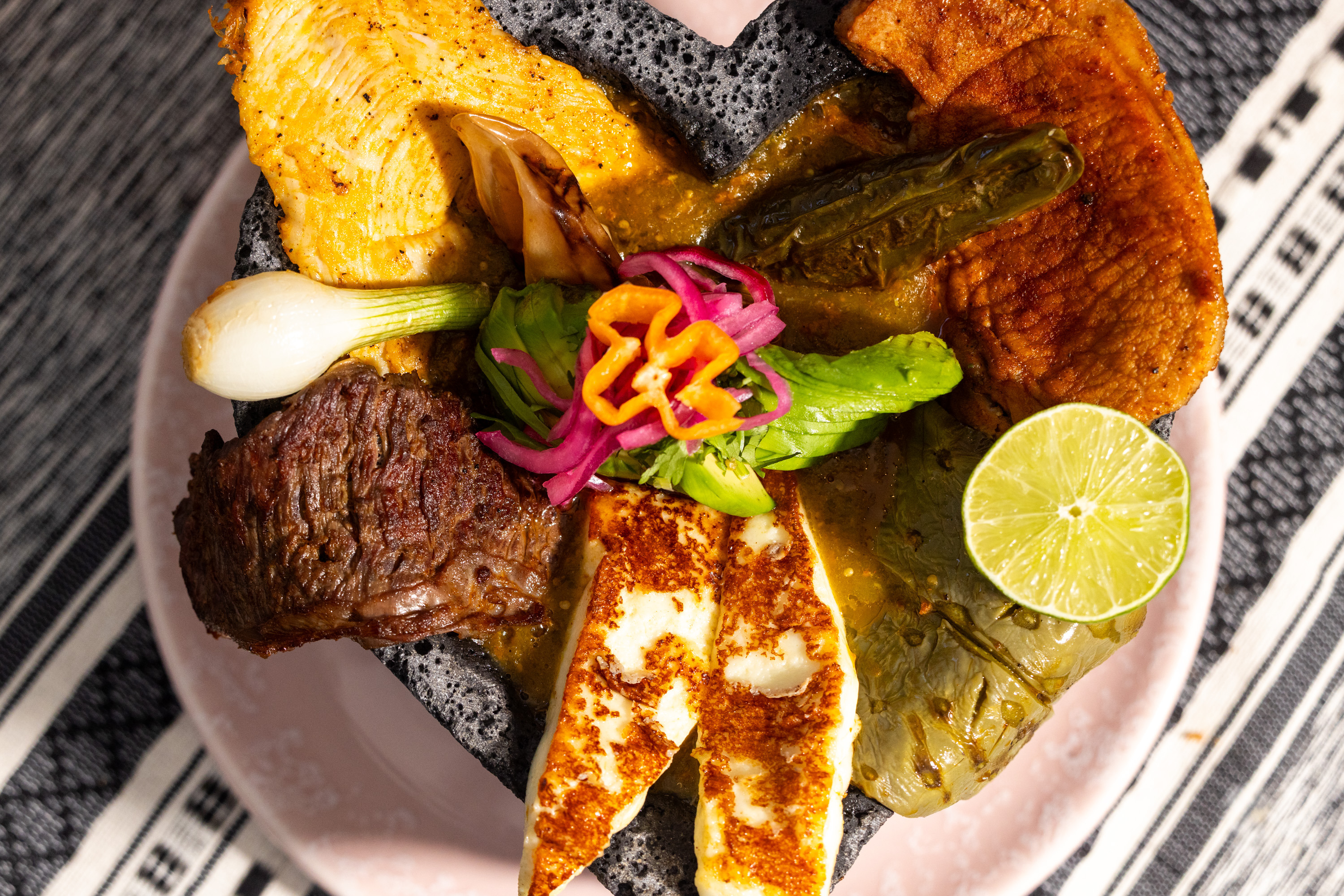A volcanic stone bowl holds grilled meats, cheese, roasted vegetables, a lime wedge, and garnished with sliced chilies and herbs, all atop a colorful sauce.