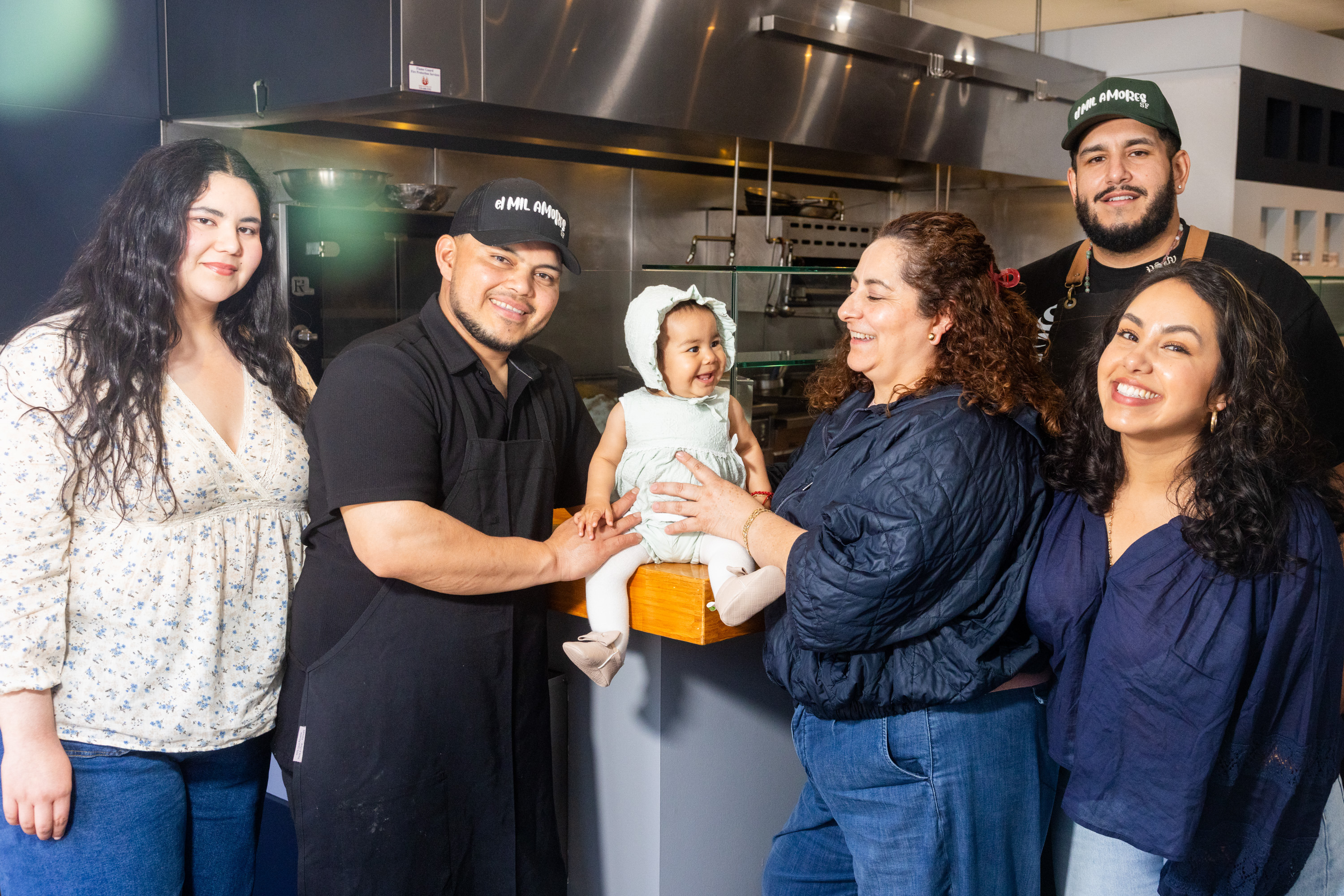 A group of six people, with one being a baby, are gathered in a kitchen setting. They are smiling and interacting warmly with each other.