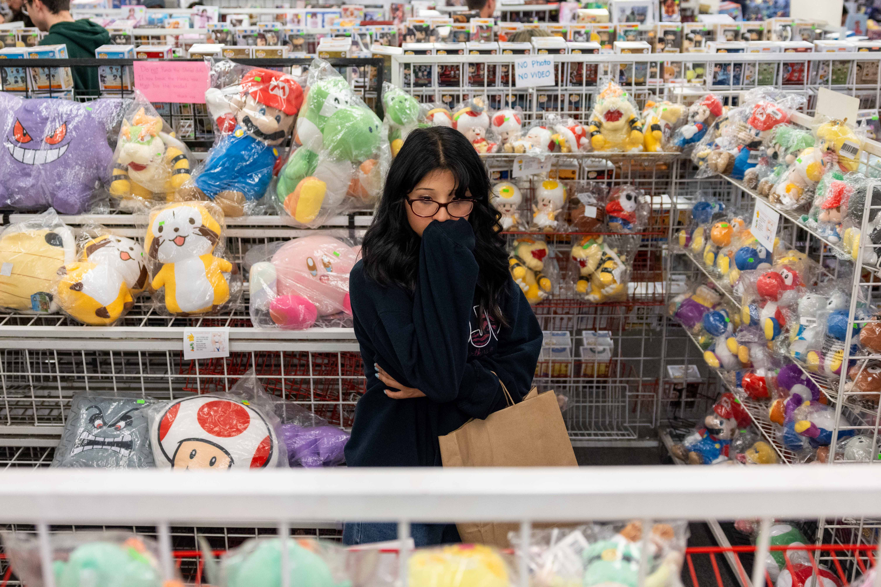 A woman with glasses stands in front of shelves filled with various plush toys, including characters from popular games. She's holding a shopping bag and appears thoughtful.
