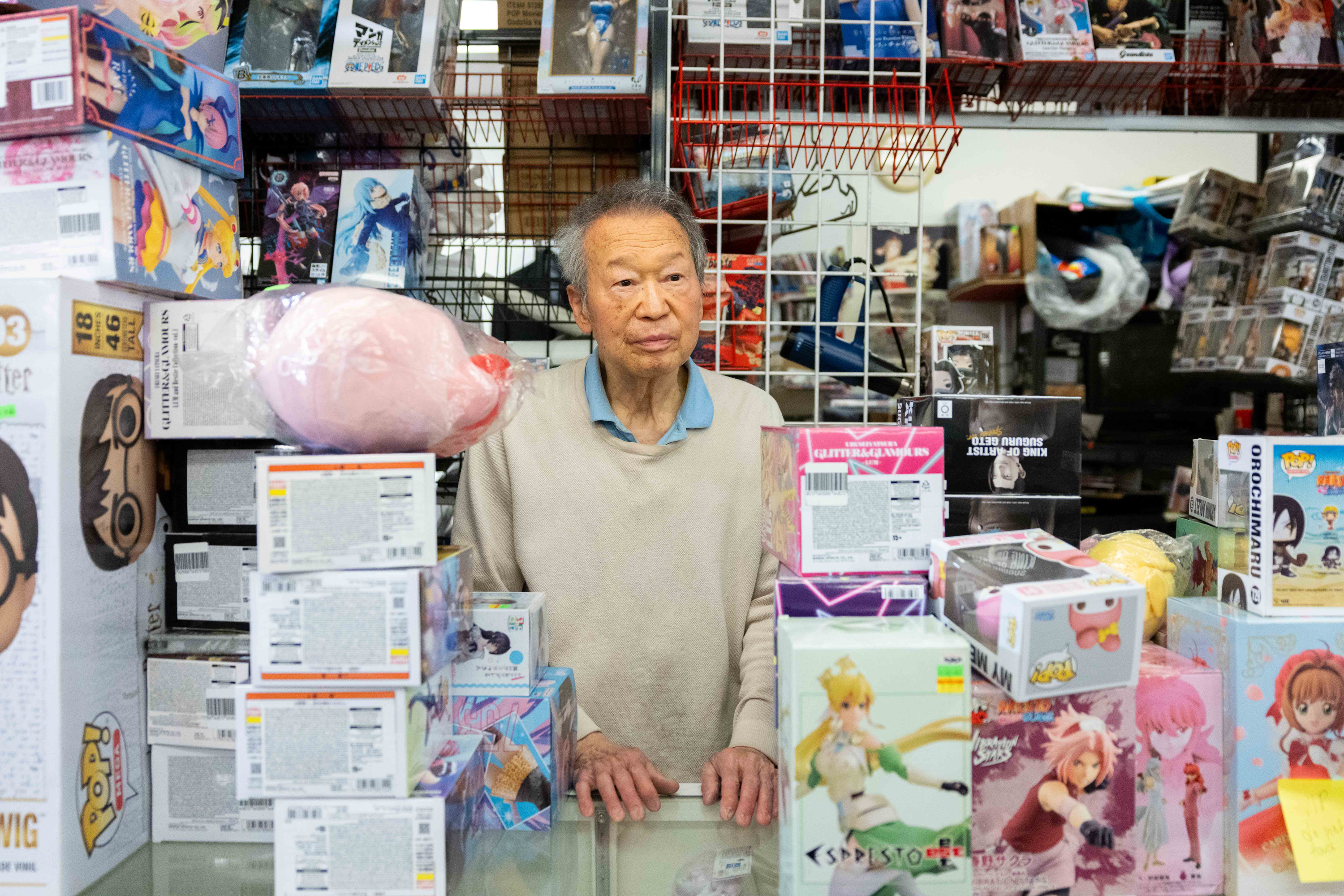 An elderly man stands behind a glass counter filled with anime figurine boxes. Shelves in the background display more collectible figures and toys.