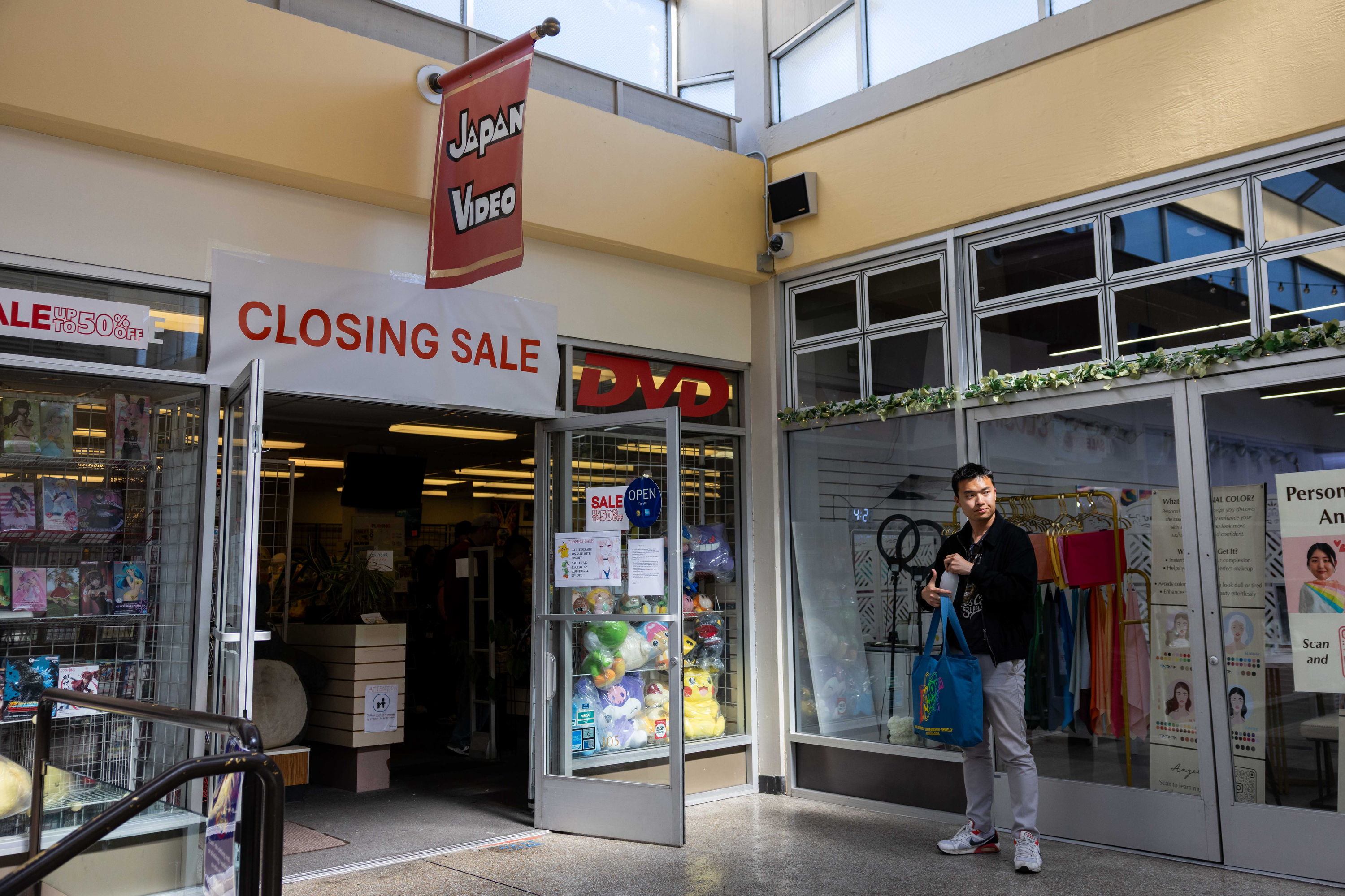 A store with a &quot;Closing Sale&quot; sign features DVDs and plush toys in its entrance. A person holding a blue bag stands outside, next to another shop with colorful items.