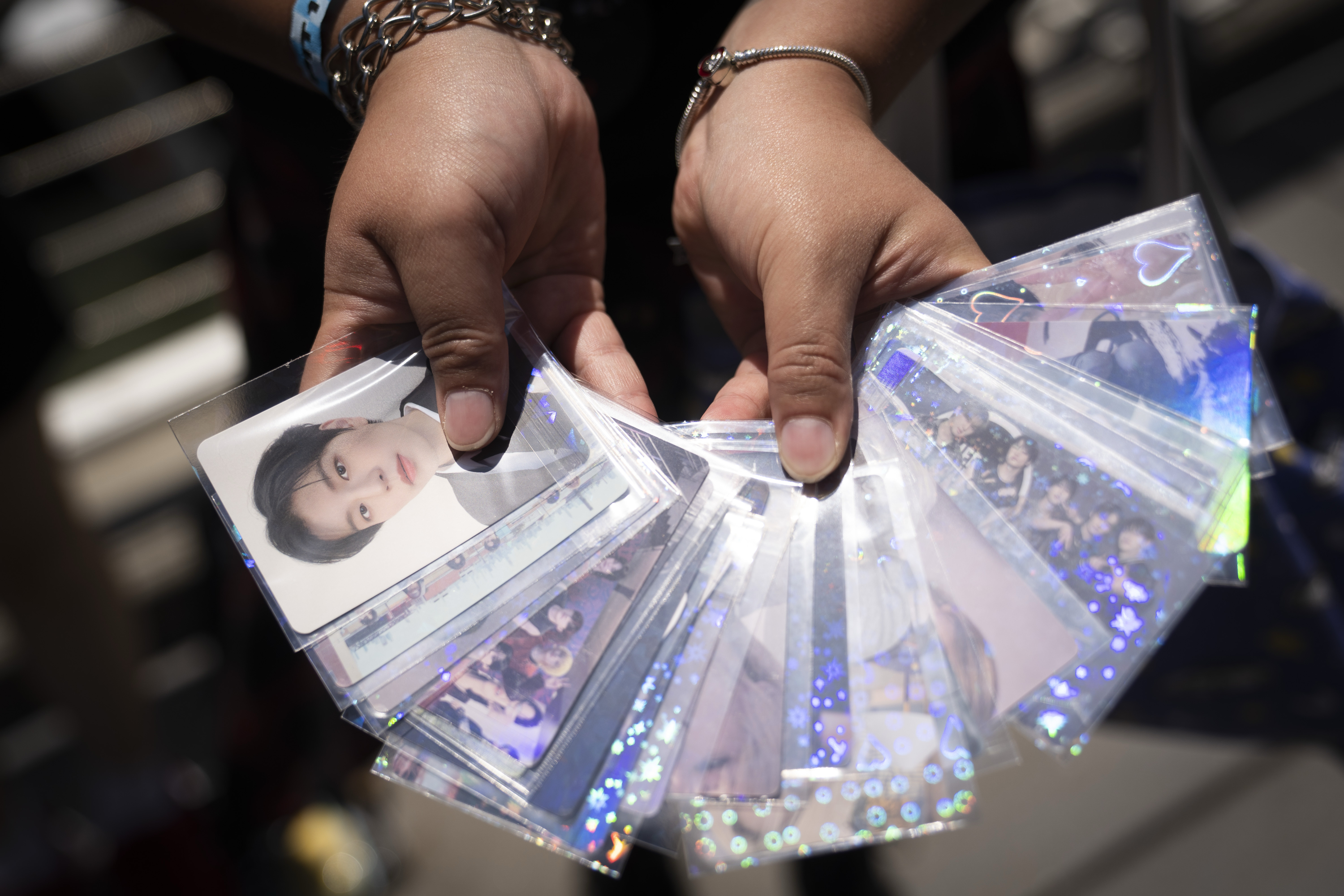 Two hands hold a fan of K-pop photo cards featuring people in sparkling sleeves. A wristband and bracelet are visible on the left wrist.