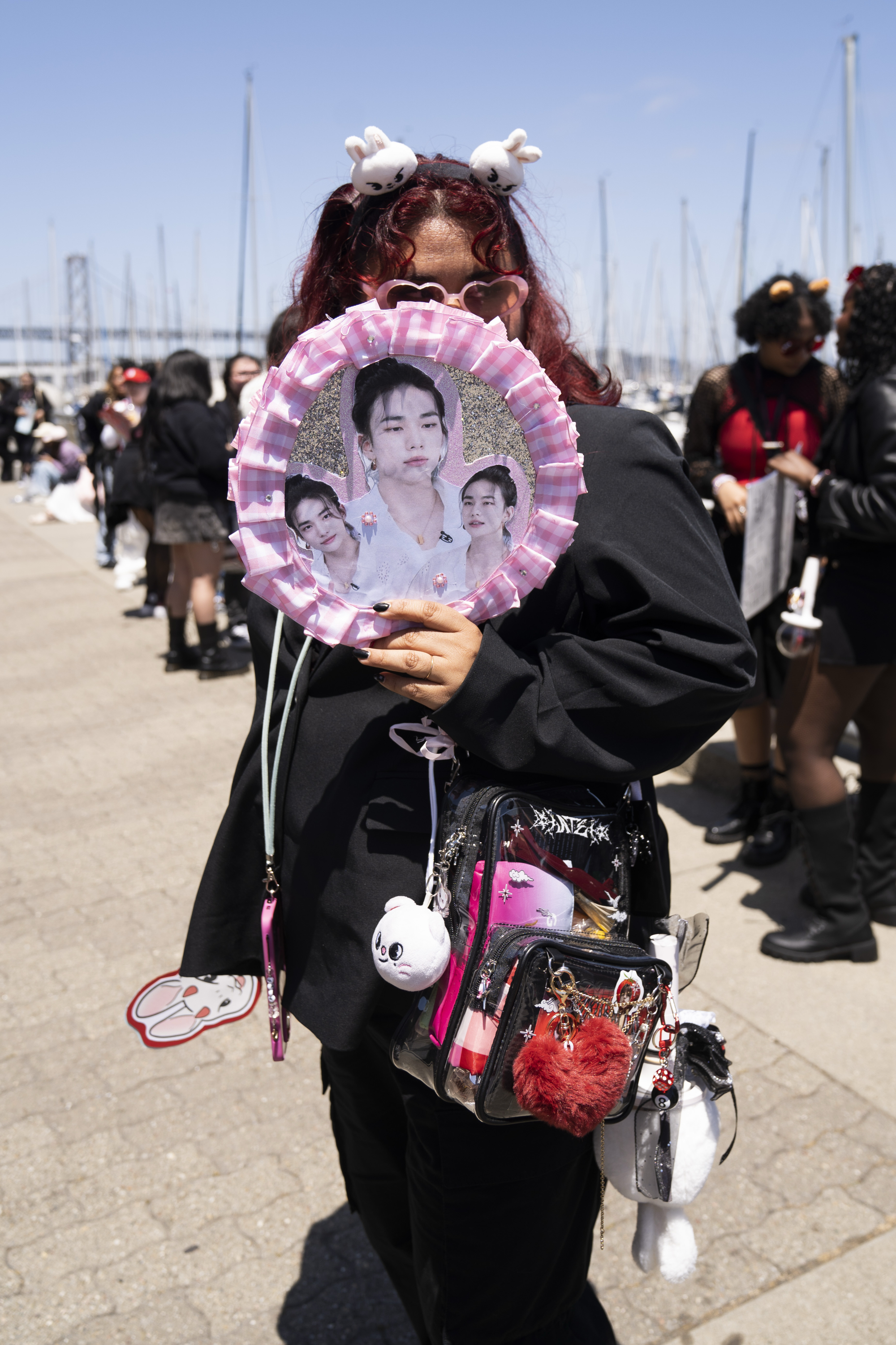 A person with red hair wears a headband, holding a pink fan with pictures. They have a clear bag filled with plushies and accessories; others stand in the background.