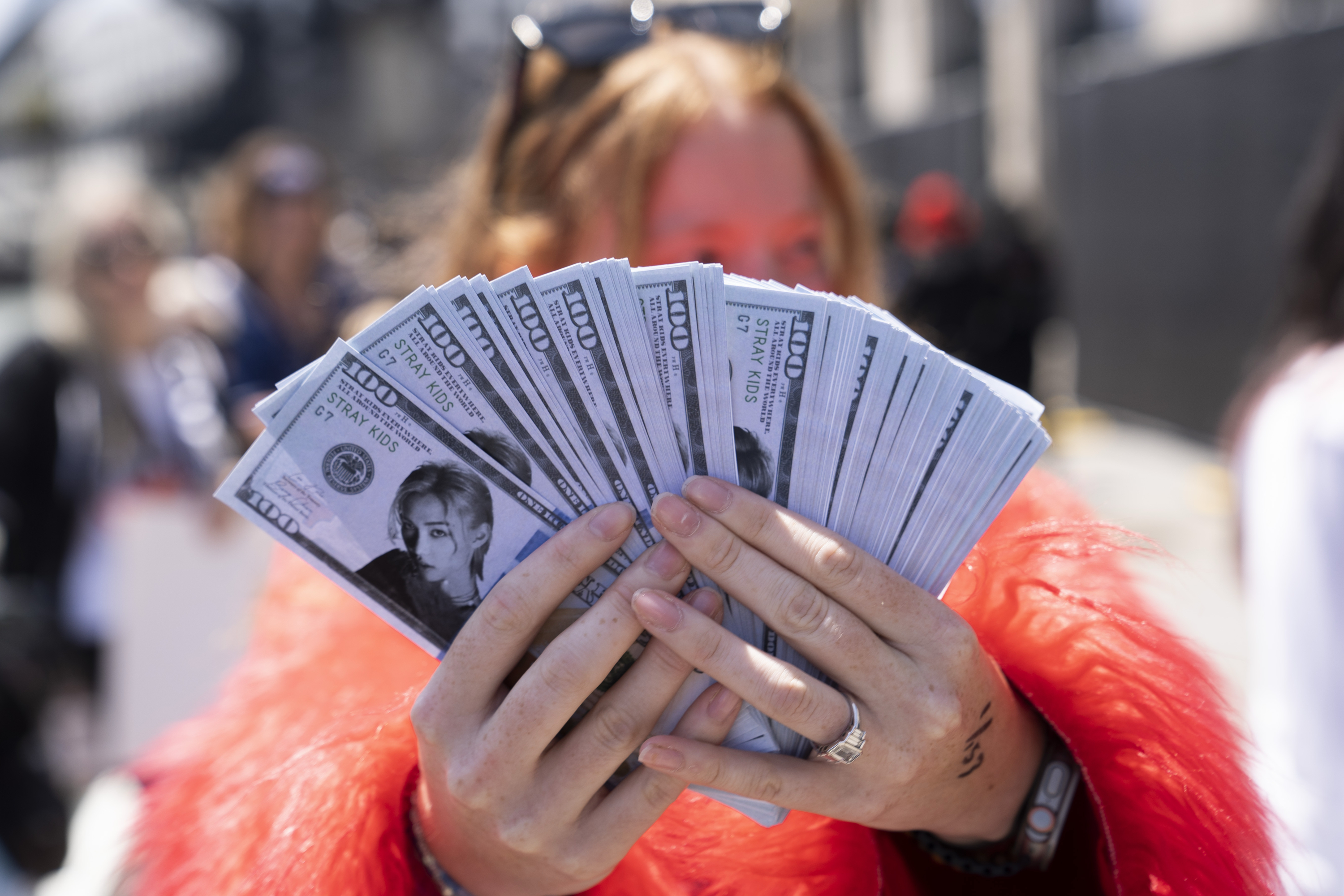A person in a red jacket holds multiple fans of novelty bills featuring a portrait labeled &quot;Stray Kids.&quot; The background is blurred with indistinct figures.