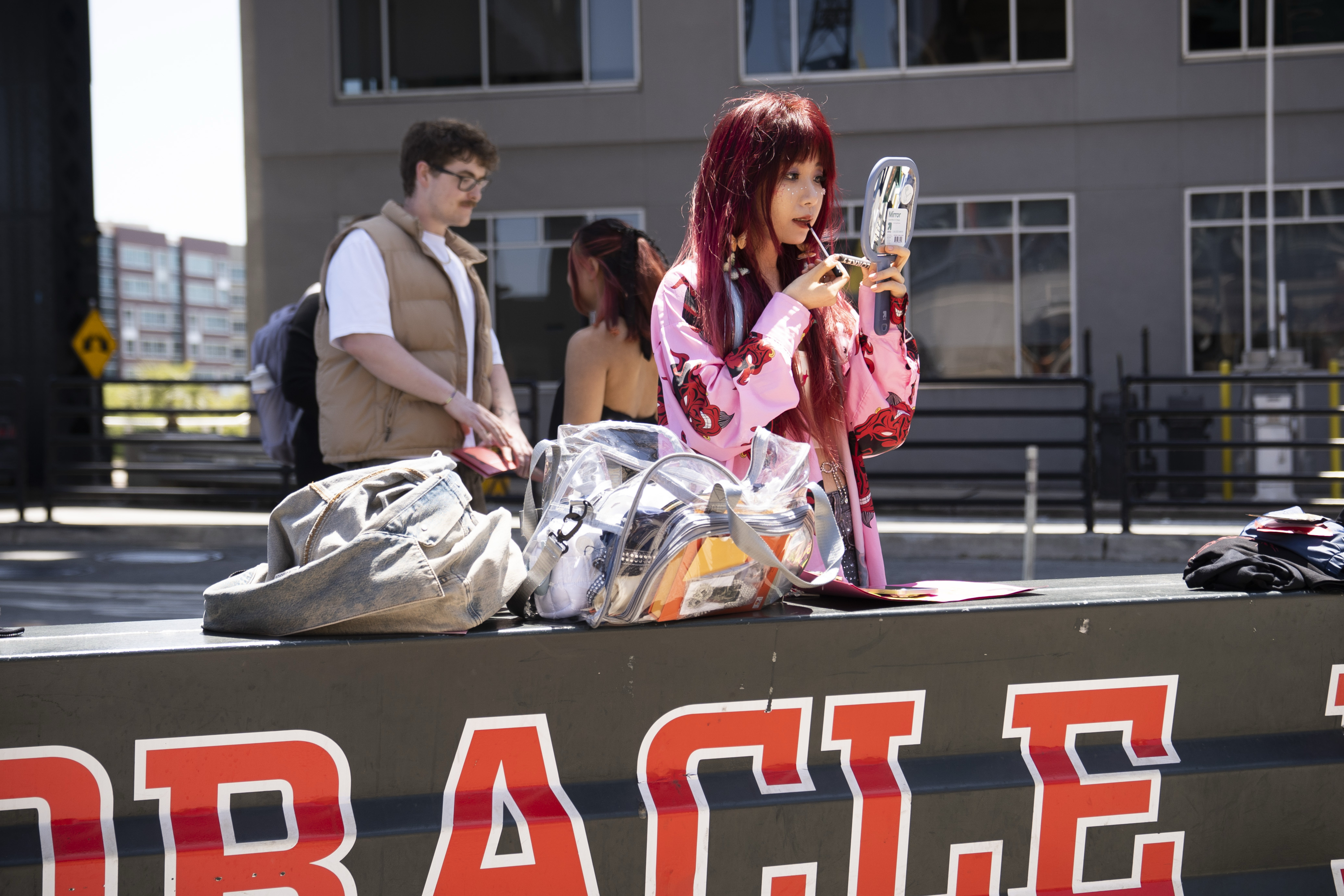 A person with long red hair in a pink outfit applies lipstick using a hand mirror. Nearby, a man stands, and bags are on a bench labeled &quot;ORACLE.&quot;