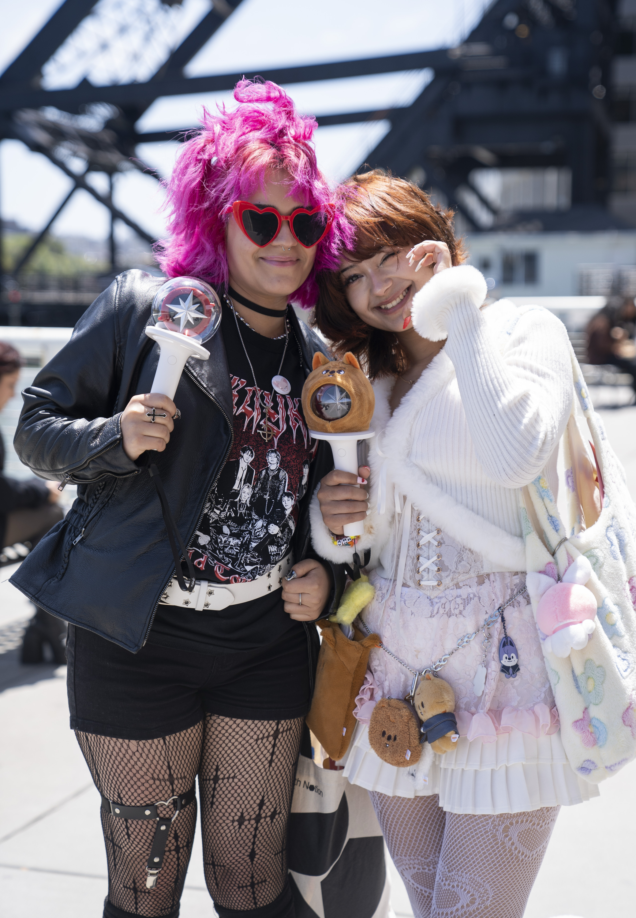 Two friends pose happily outdoors. One has bright pink hair, heart-shaped sunglasses, and a leather jacket. The other wears a fluffy white outfit and lace details.