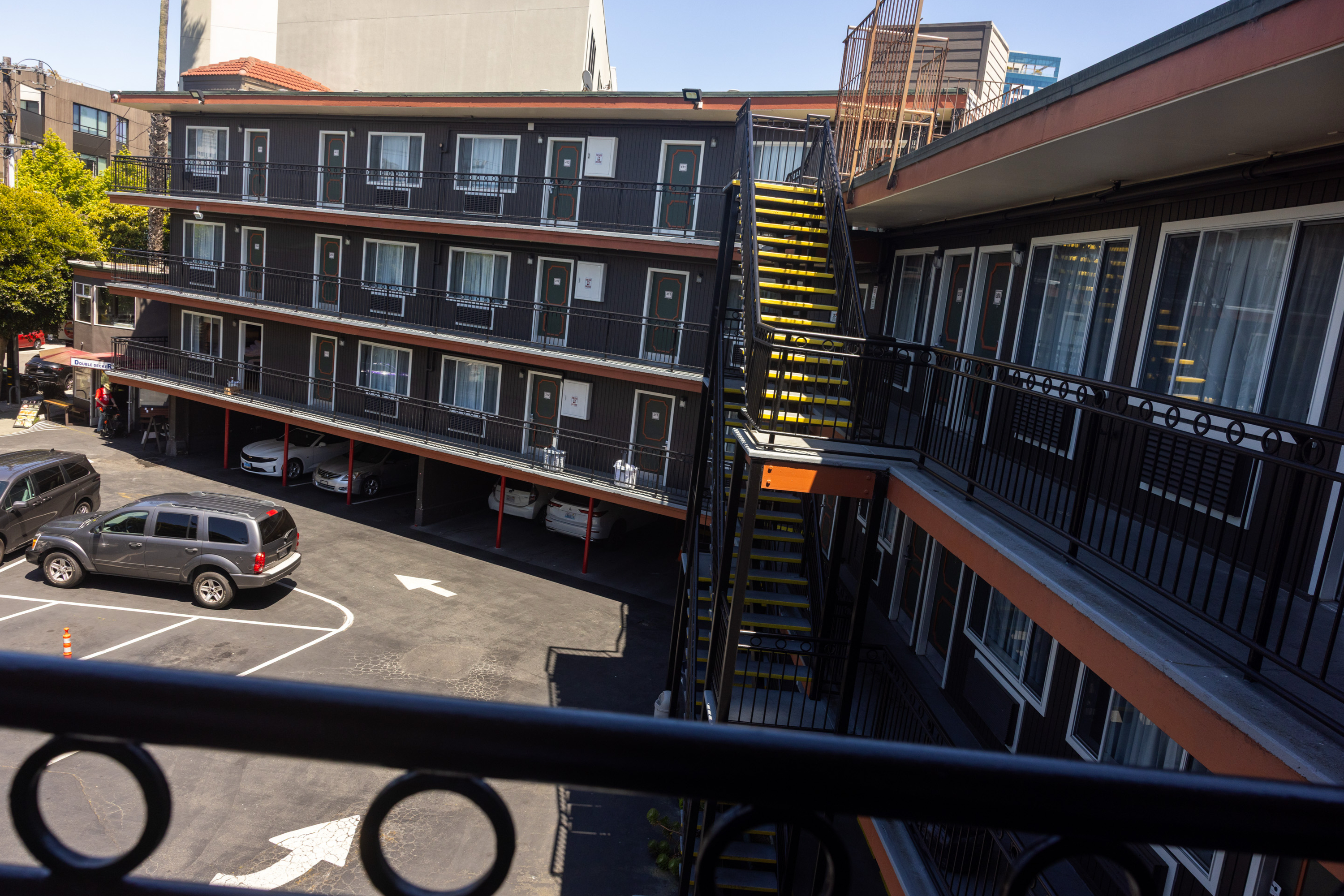 The image shows a three-story motel with balconies and a bright yellow staircase. Below, cars are parked in marked spaces on a paved lot.