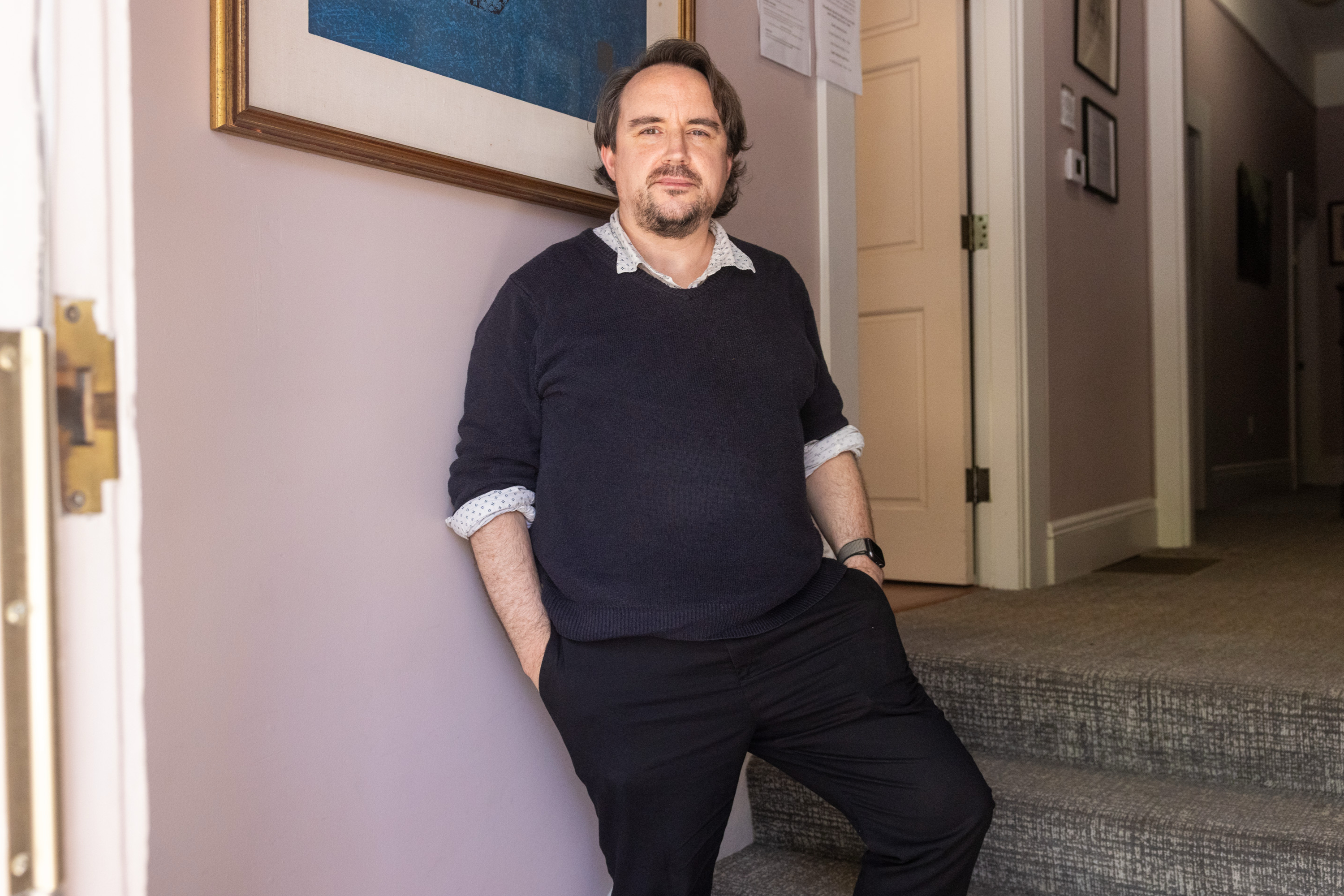 A man with dark hair and a beard leans against a wall in a hallway, wearing a dark sweater and trousers, with one hand in his pocket. There's artwork on the wall.