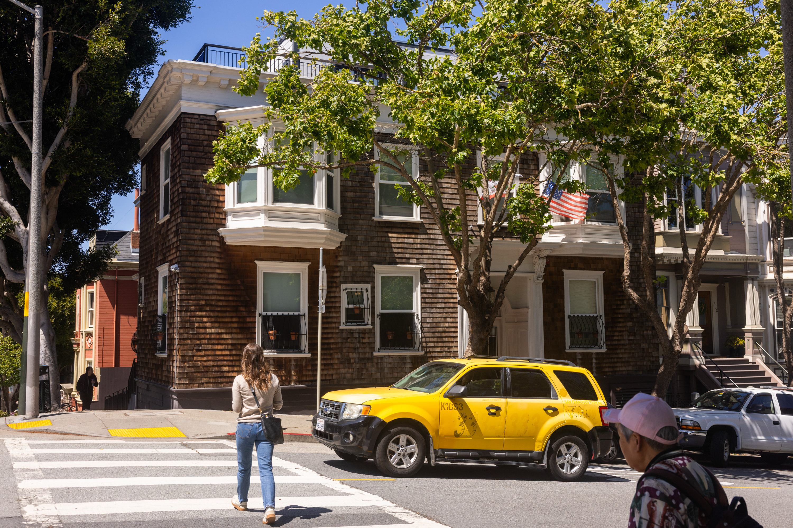 A woman crosses a street towards a corner house with wooden siding and a tree in front. A yellow SUV is parked nearby, and an American flag hangs from the house.