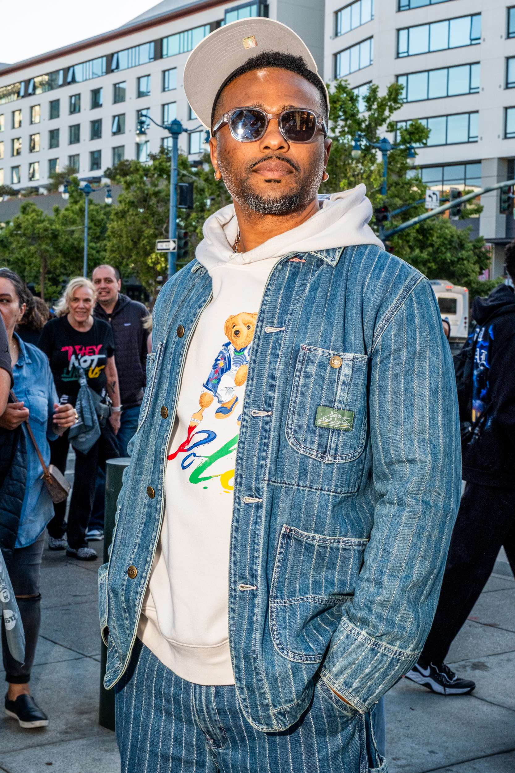 A man in a denim jacket and pants with a printed hoodie featuring a bear stands on a city sidewalk. He's wearing sunglasses and a cap, with people in the background.