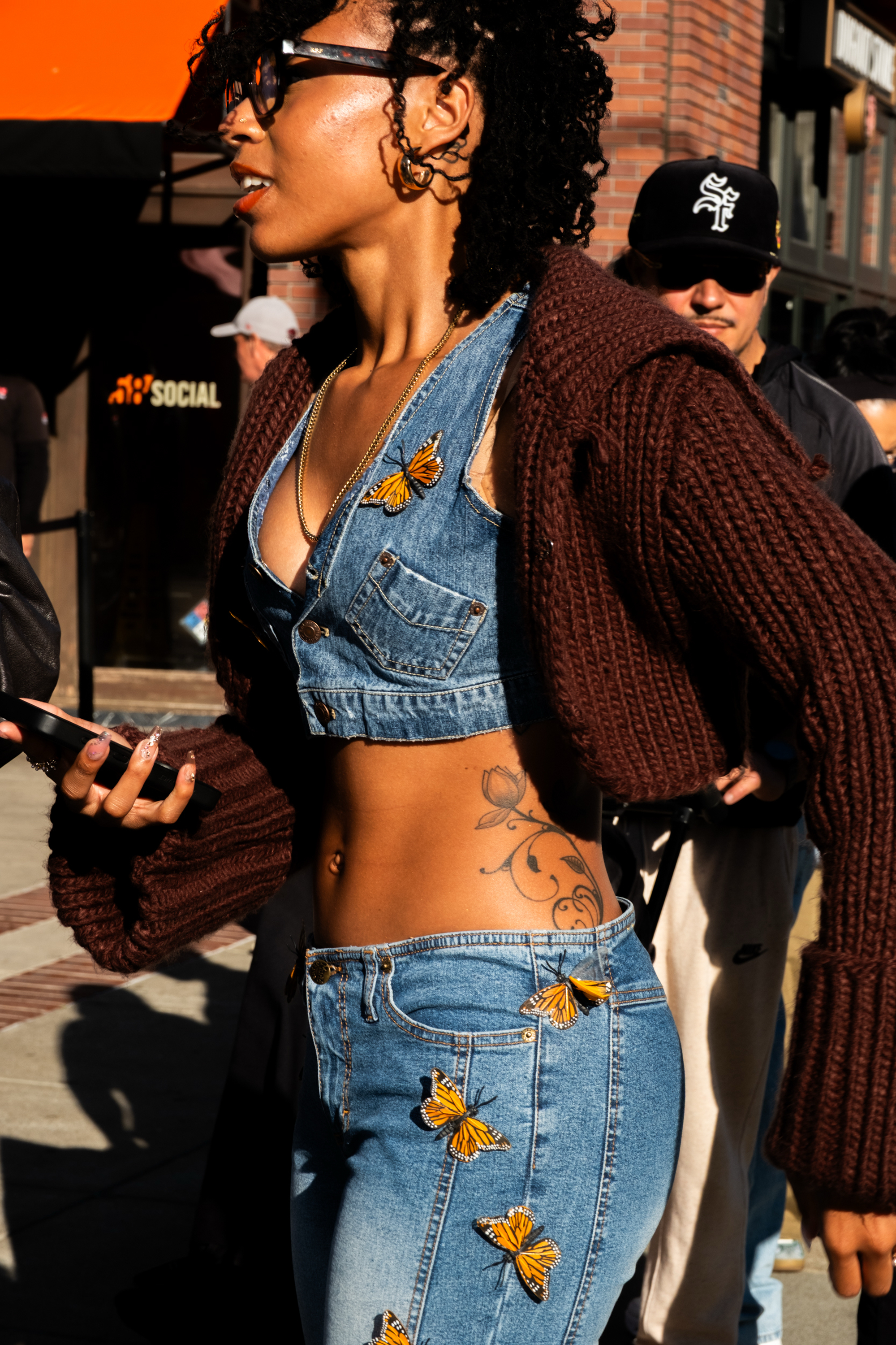 A person in sunglasses wears a denim outfit adorned with butterfly patches, a brown knitted sweater, and sports a floral tattoo. They hold a phone while walking.