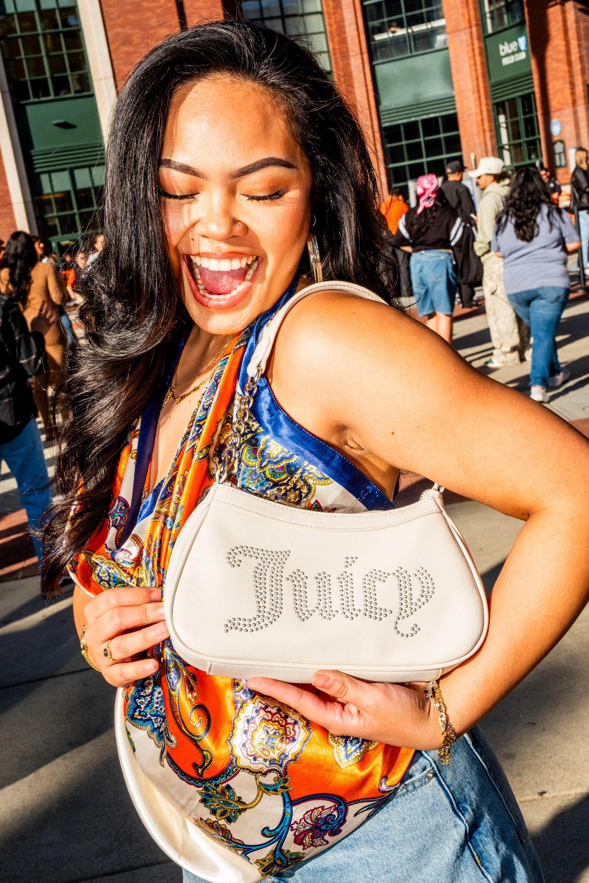 A woman smiles with closed eyes, holding a white bag labeled &quot;Juicy&quot; in rhinestones. She's wearing colorful patterned clothing and standing outdoors.