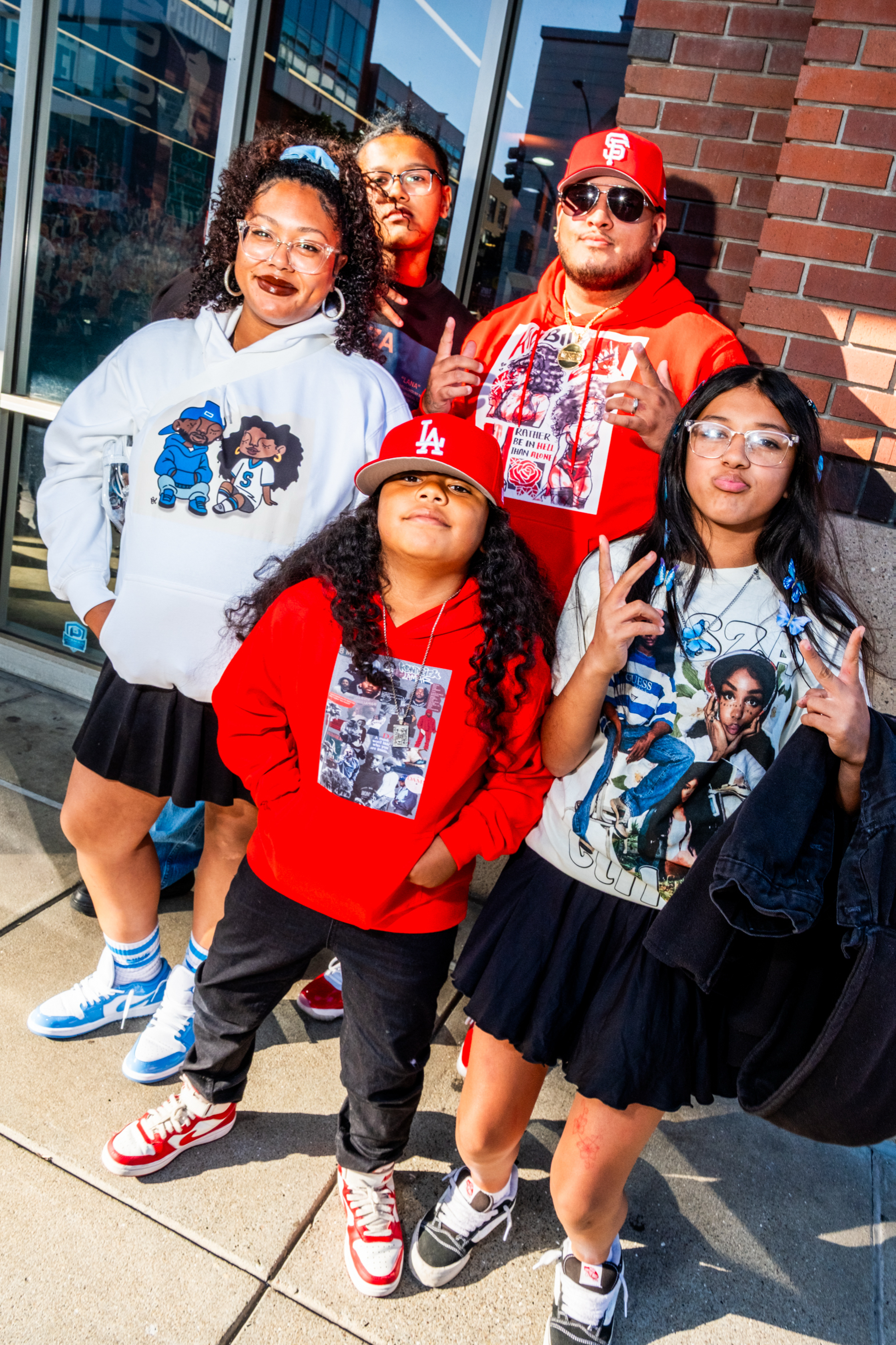 Five people pose in colorful outfits on a city sidewalk. They wear sneakers, hoodies, and graphic tees, with two making peace signs. They're smiling confidently.