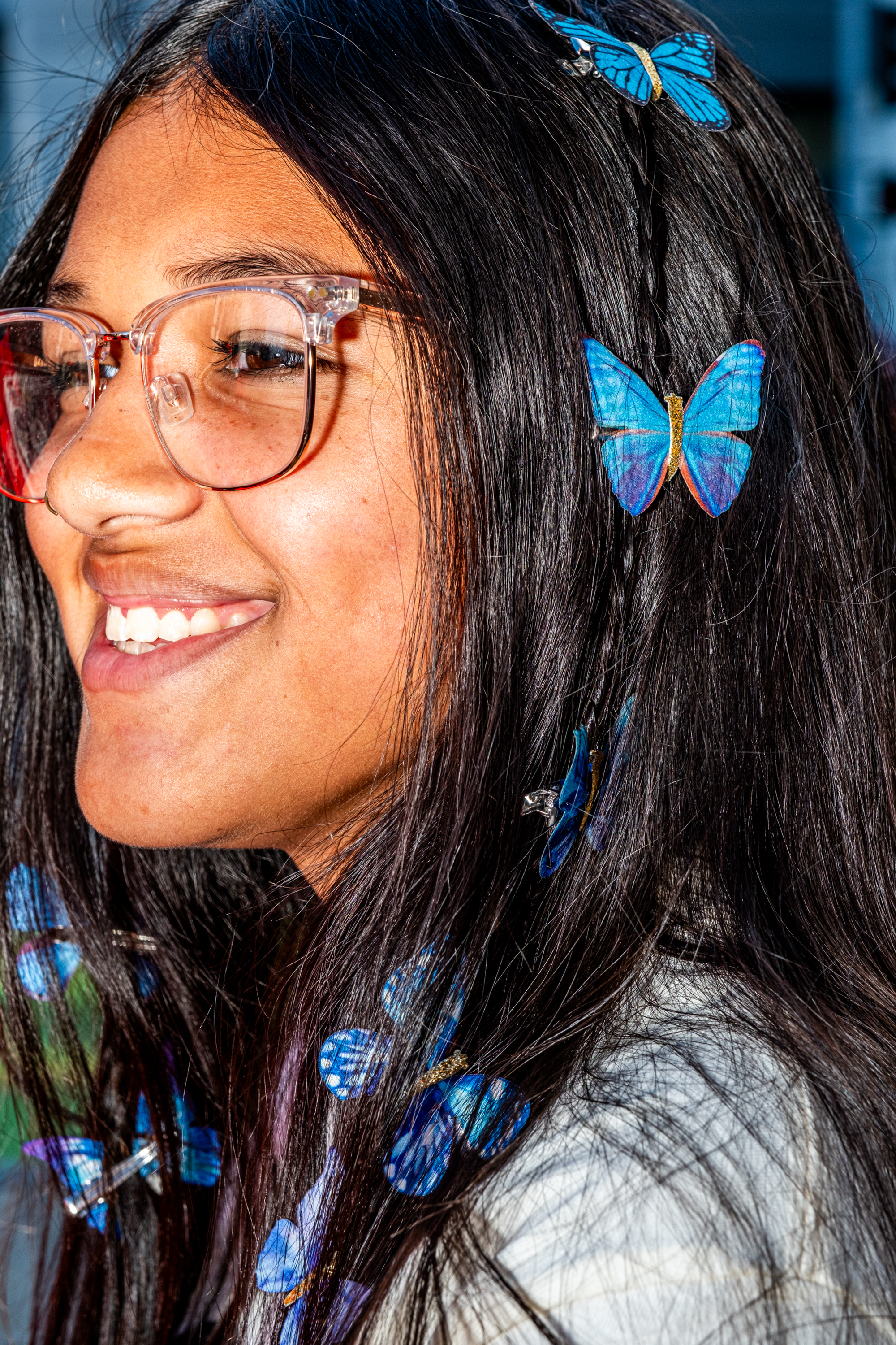 A person smiles warmly with long dark hair adorned with blue butterfly clips, wearing clear glasses and a nose ring.