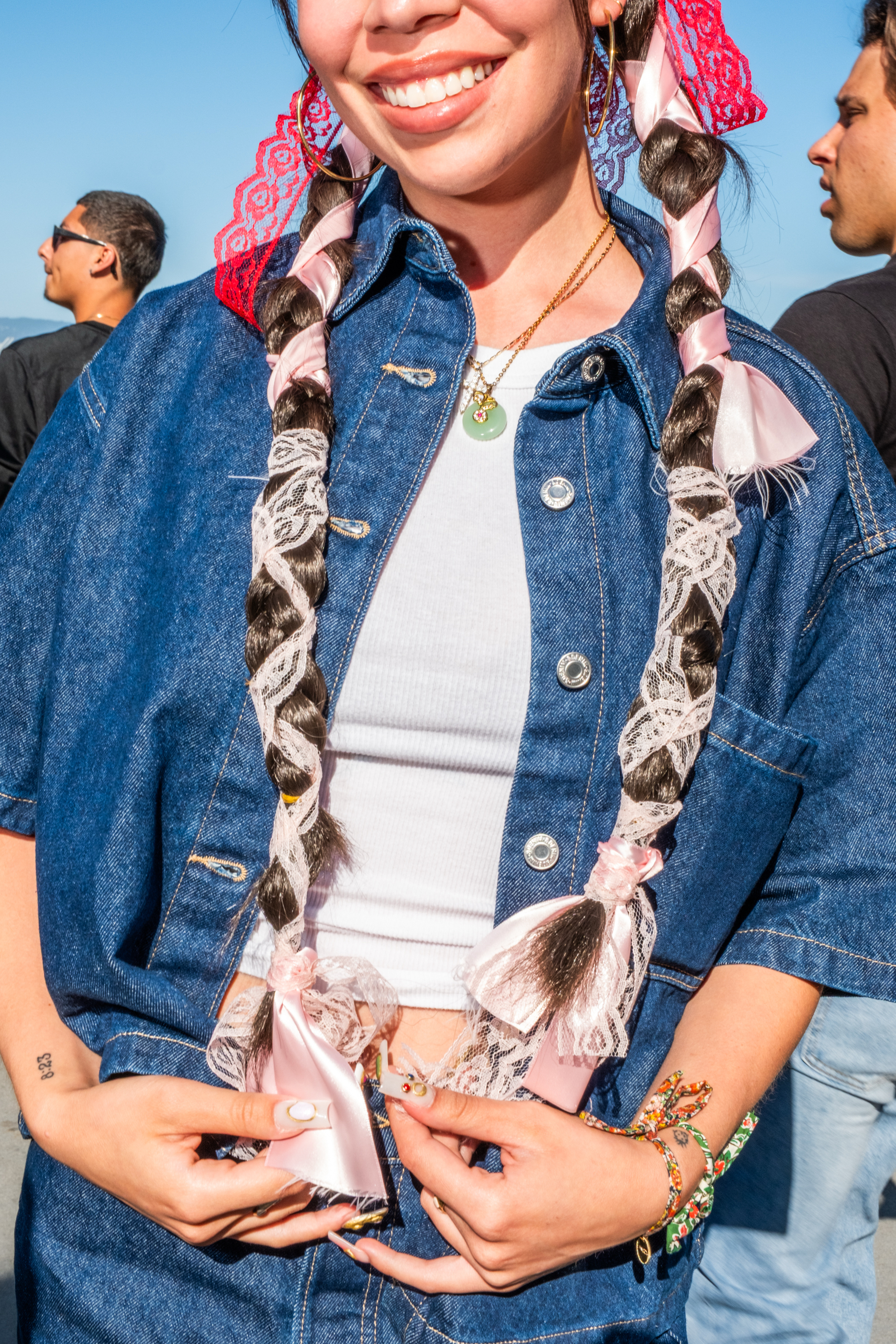 A smiling person with long braids wrapped in pink ribbon and lace, wearing a denim jacket over a white top, accessorized with necklaces and bracelets.
