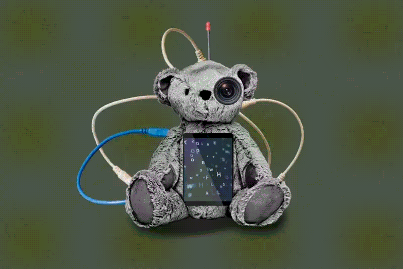 A teddy bear with one eye replaced by a camera lens sits with a screen showing letters on its belly. Various cables are connected to it.