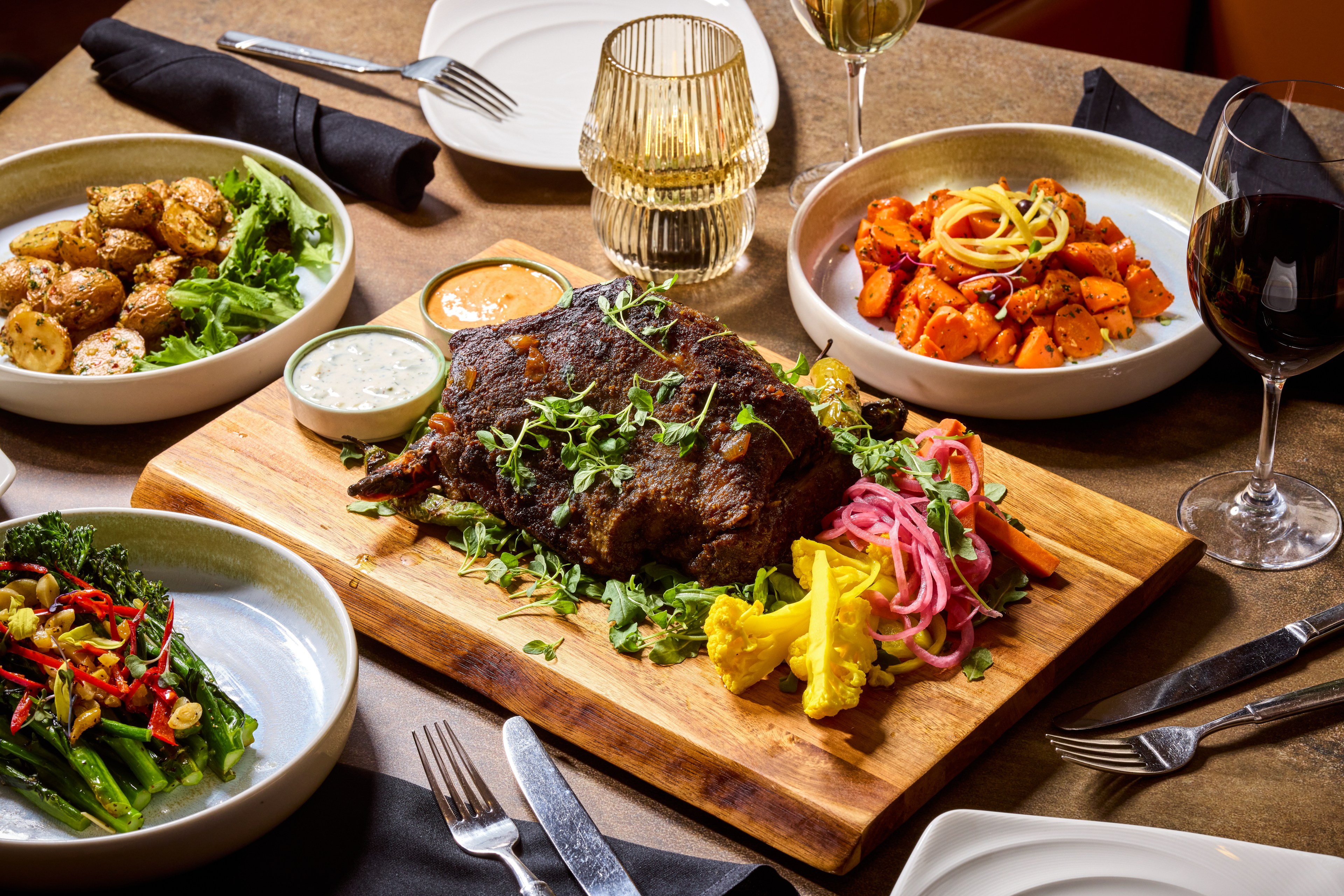 A wooden board with roasted meat garnished with herbs and colorful pickled vegetables, surrounded by bowls of potatoes, carrots, and greens, alongside drinks.