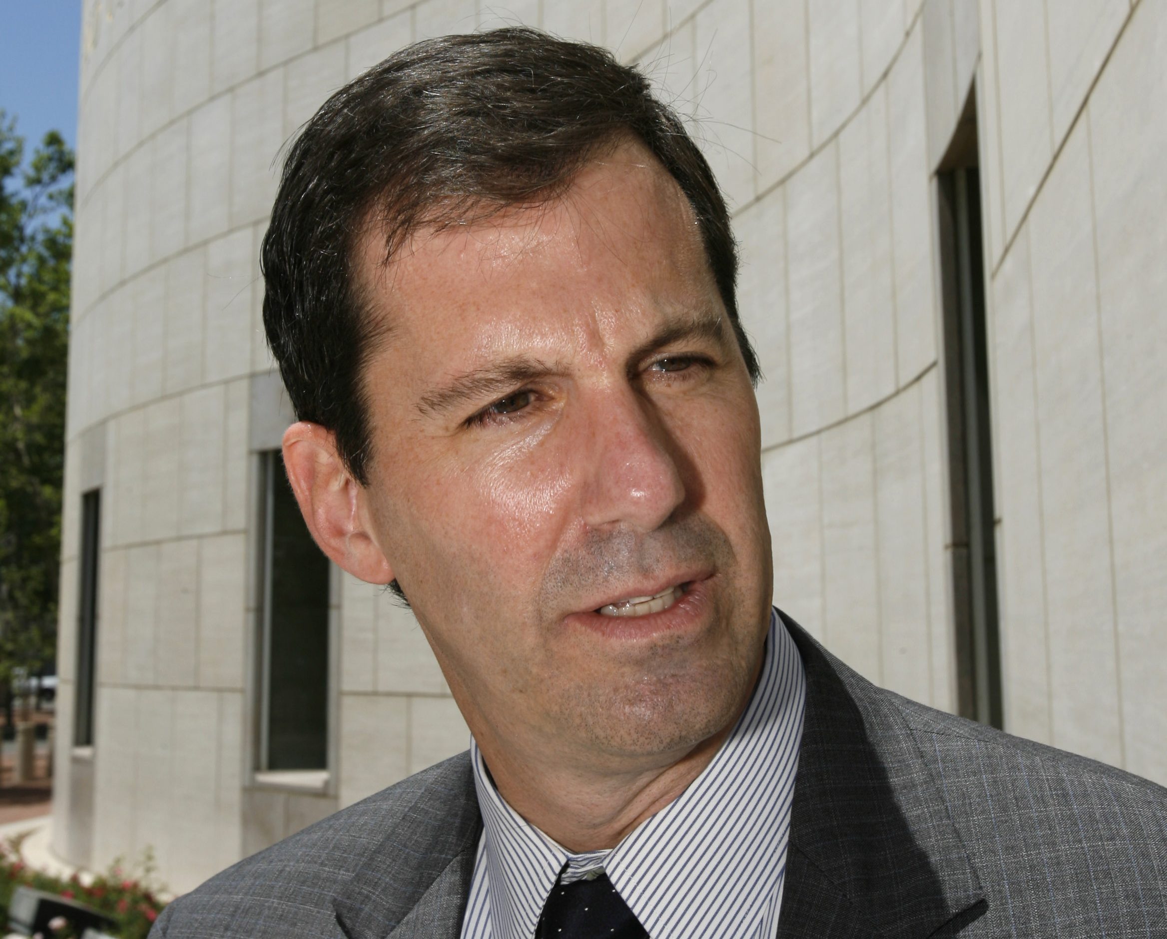 A man in a suit and tie stands outside, looking to the side. The background shows a curved, light-colored building wall with windows.