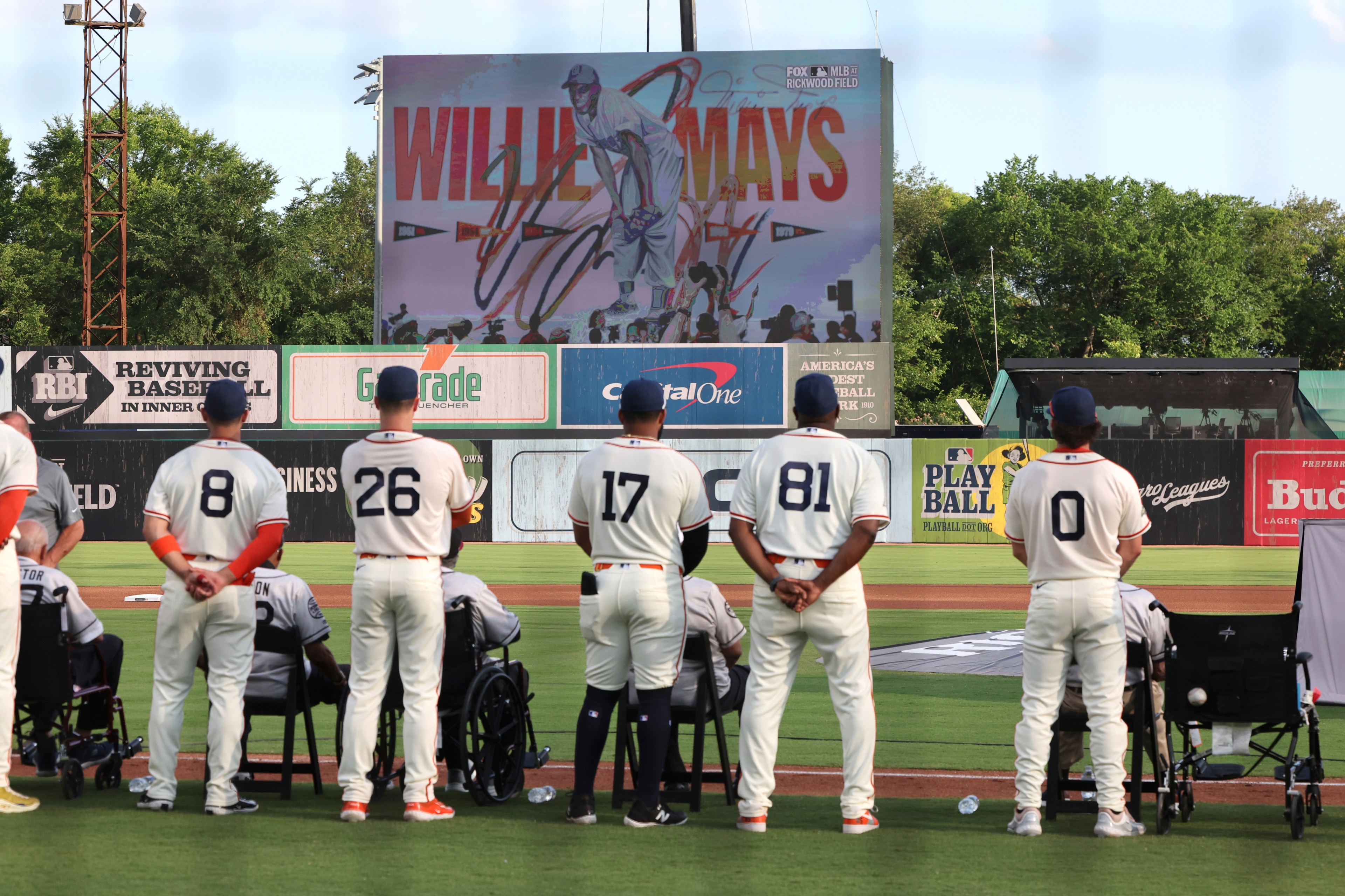Baseball players in white jerseys stand on a field, facing a giant screen displaying &quot;Willie Mays&quot; and his image. Some individuals are seated in wheelchairs.