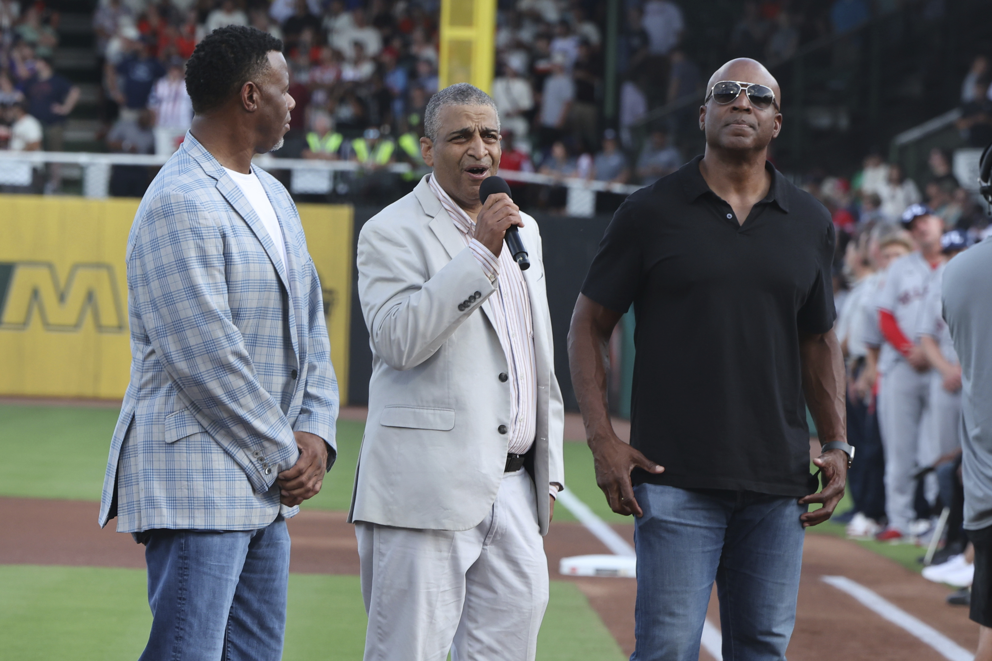 Three men stand on a baseball field. One wears a plaid blazer, another a light suit singing into a microphone, and the third in a black shirt and sunglasses.