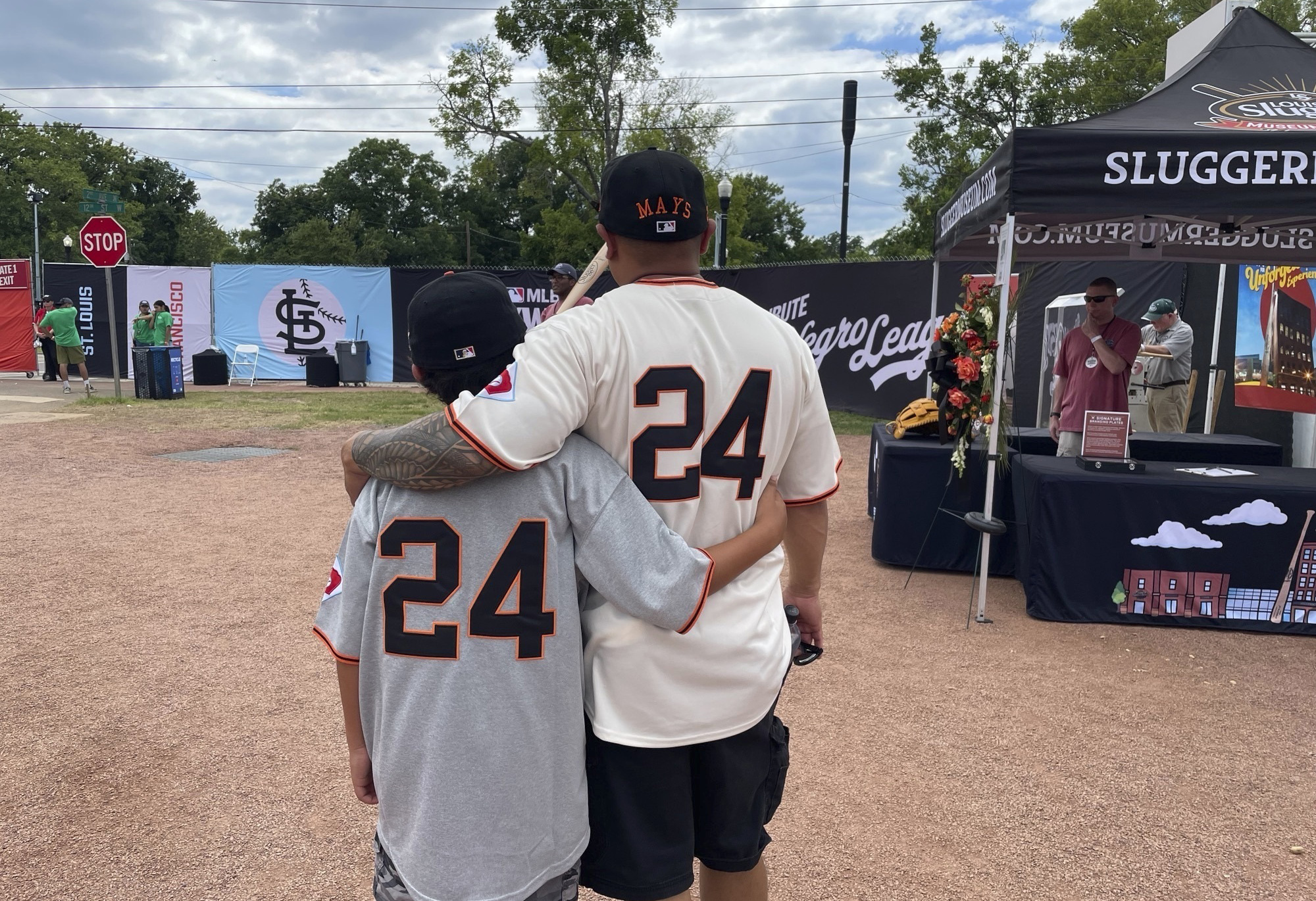 A man and a boy, wearing matching baseball jerseys with the number 24, embrace near a tent at a baseball event.