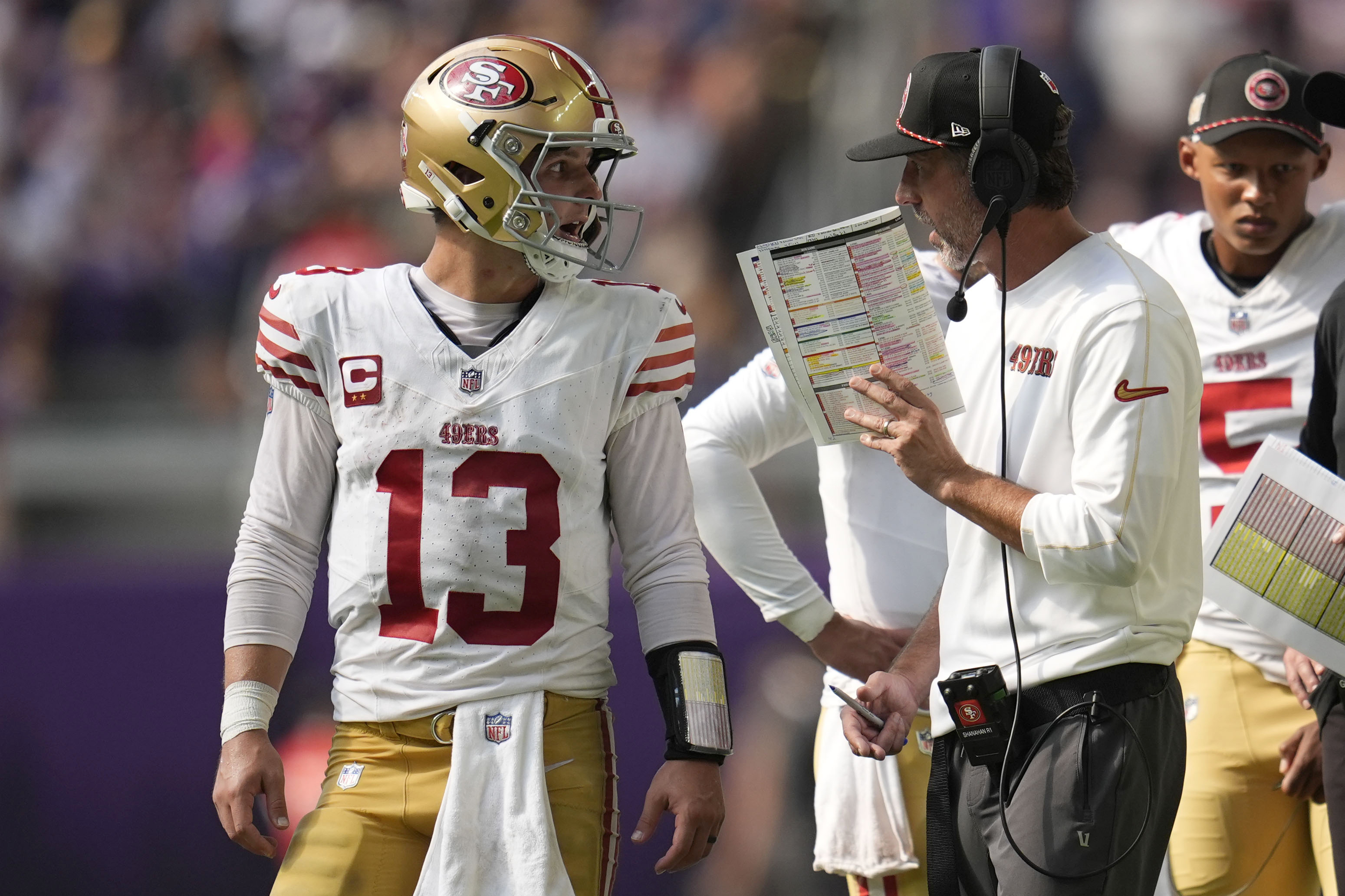 A football player in a white jersey and gold pants with a helmet listens to a coach holding a play sheet. Another person in uniform is in the background.
