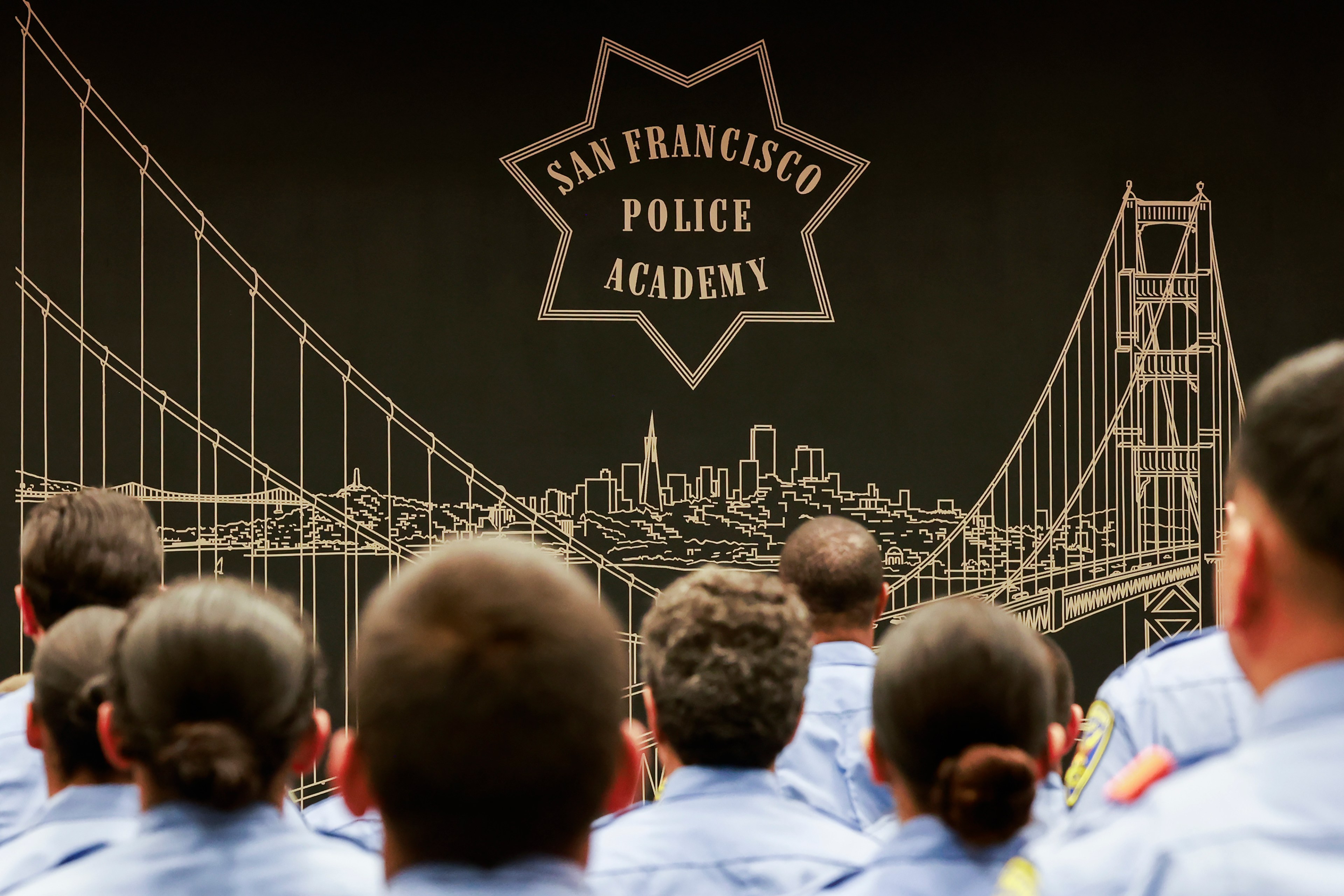 A group of uniformed individuals sits facing a large wall mural of the San Francisco skyline and bridge, with "San Francisco Police Academy" prominently displayed.