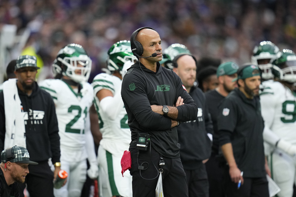 A football coach wearing a headset stands on the sideline with arms crossed. Players in green and white uniforms, also stand in the background.