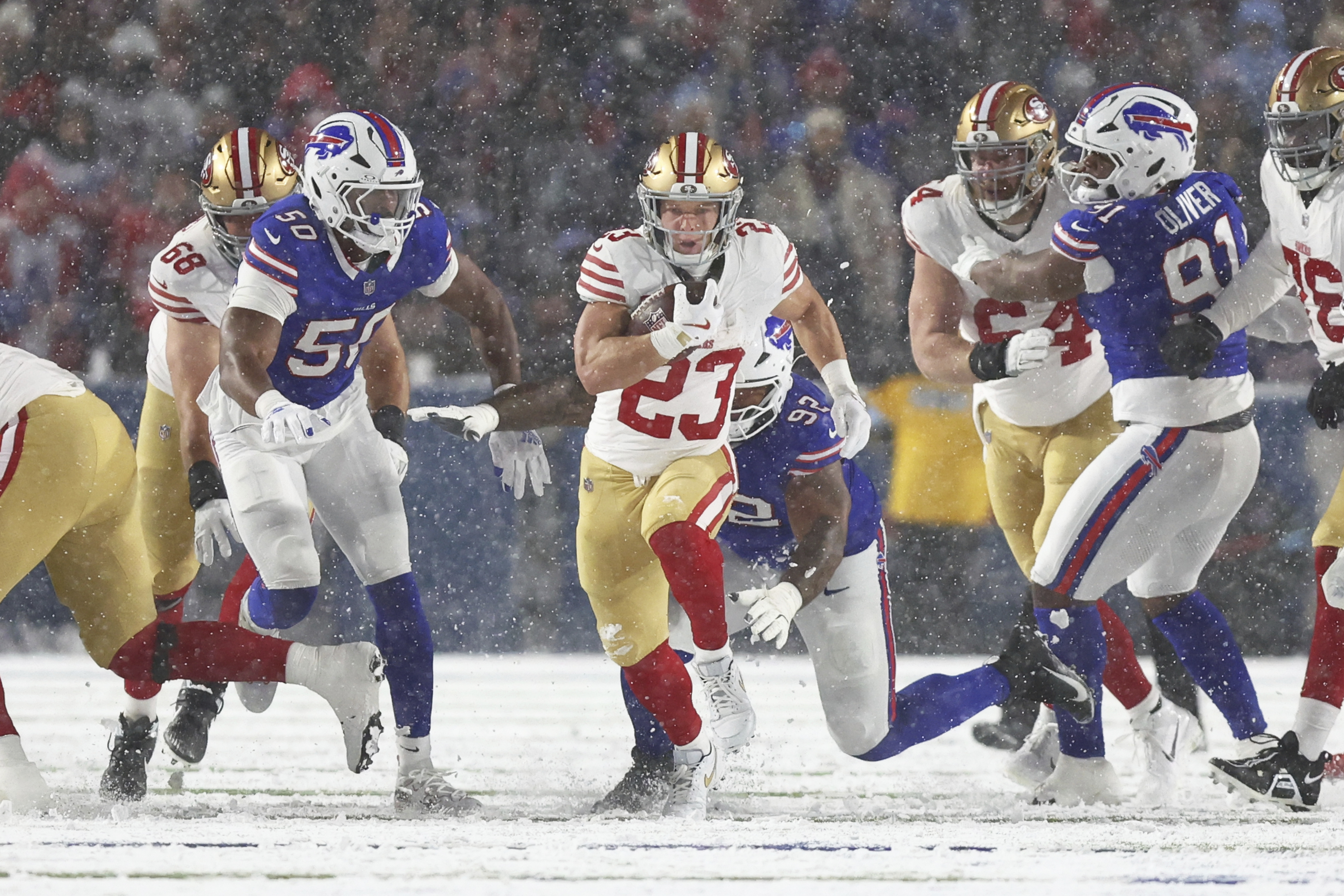 A football player in a red and gold uniform runs with the ball through snow, while players in blue and white uniforms pursue from around him.