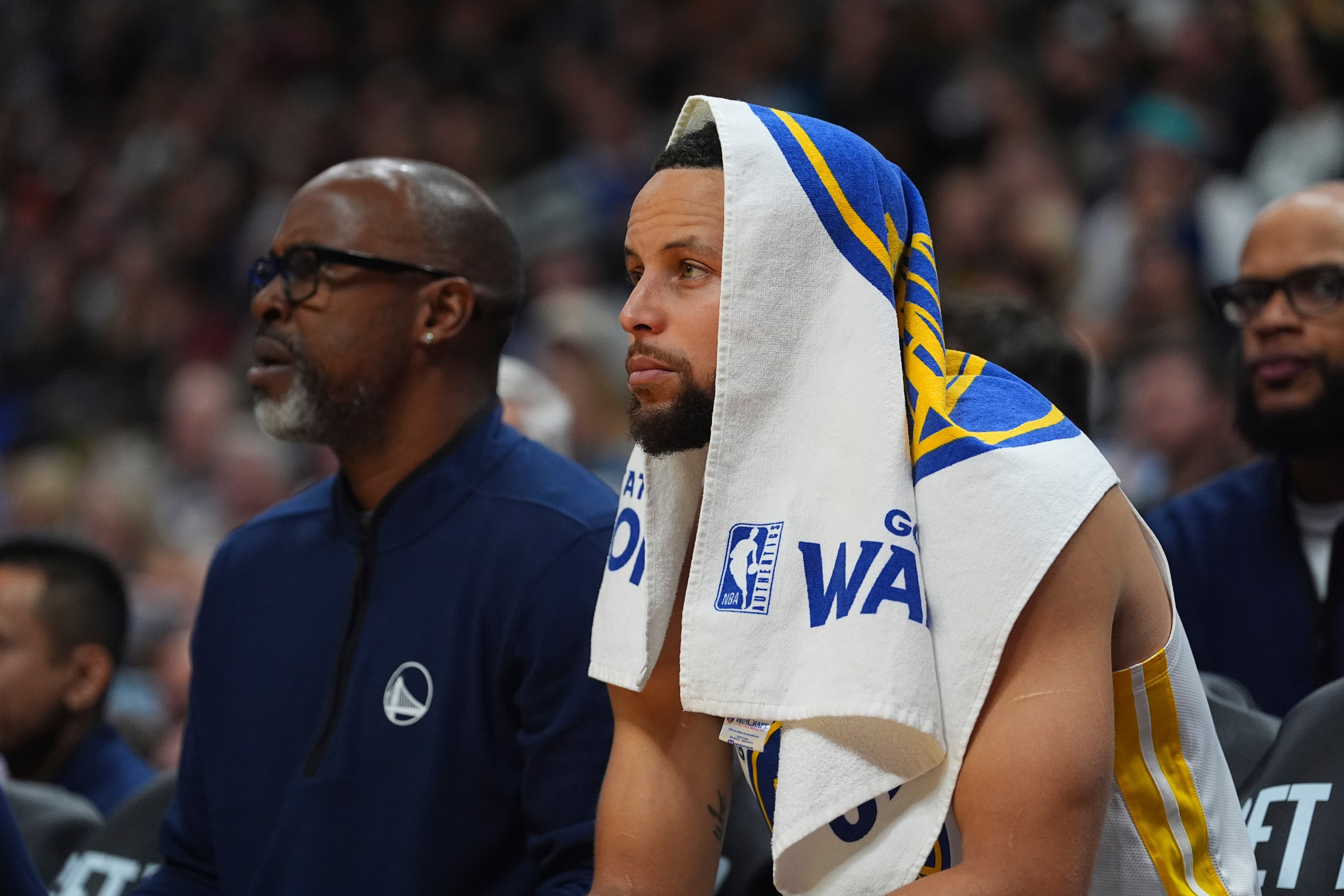 A basketball player sits on the bench with a towel draped over their head. Another person in sports attire sits beside them. The setting appears to be a game.