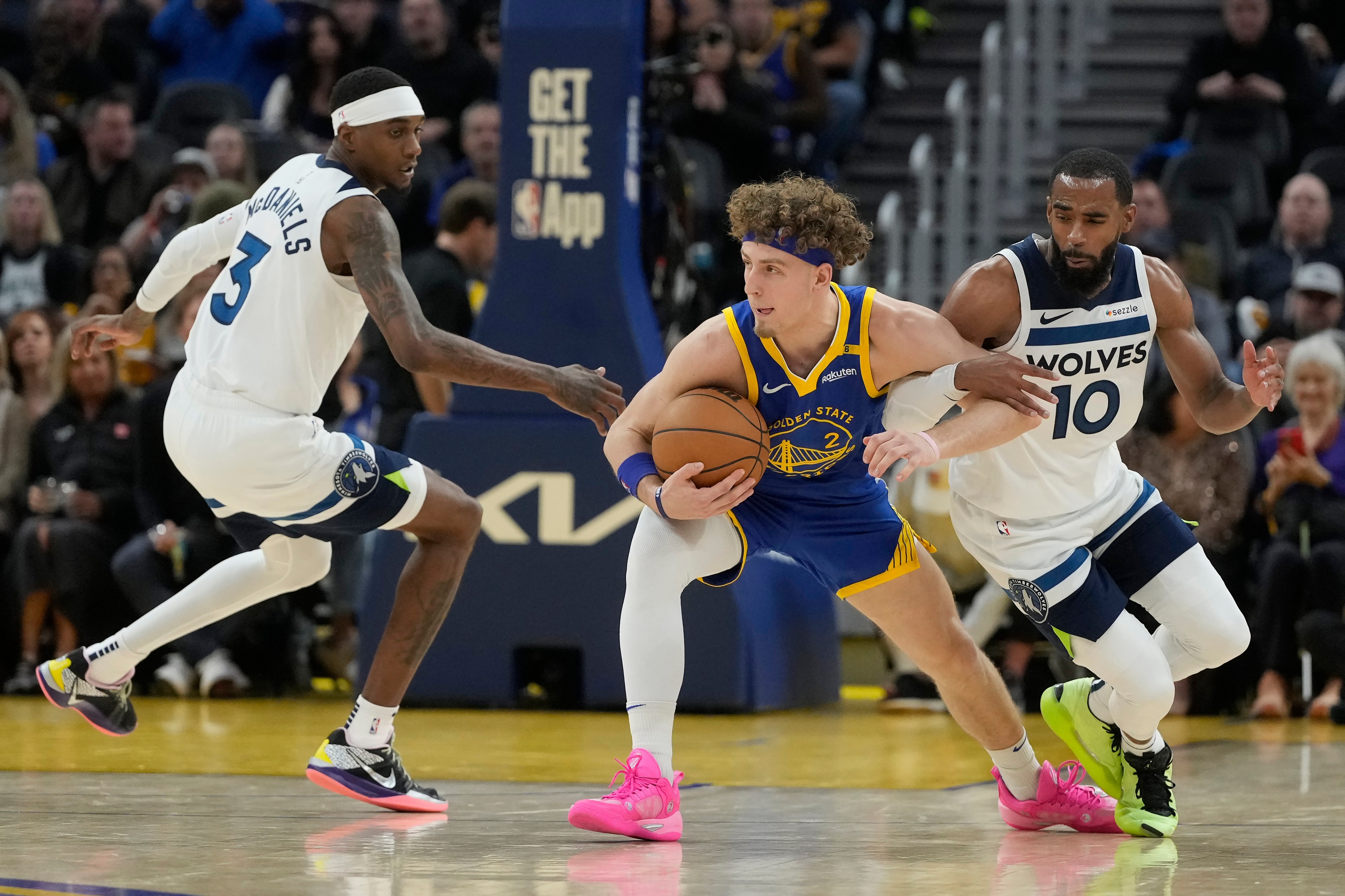 A basketball player in a blue and yellow uniform dribbles the ball while two players in white uniforms defend him on the court, with spectators in the background.
