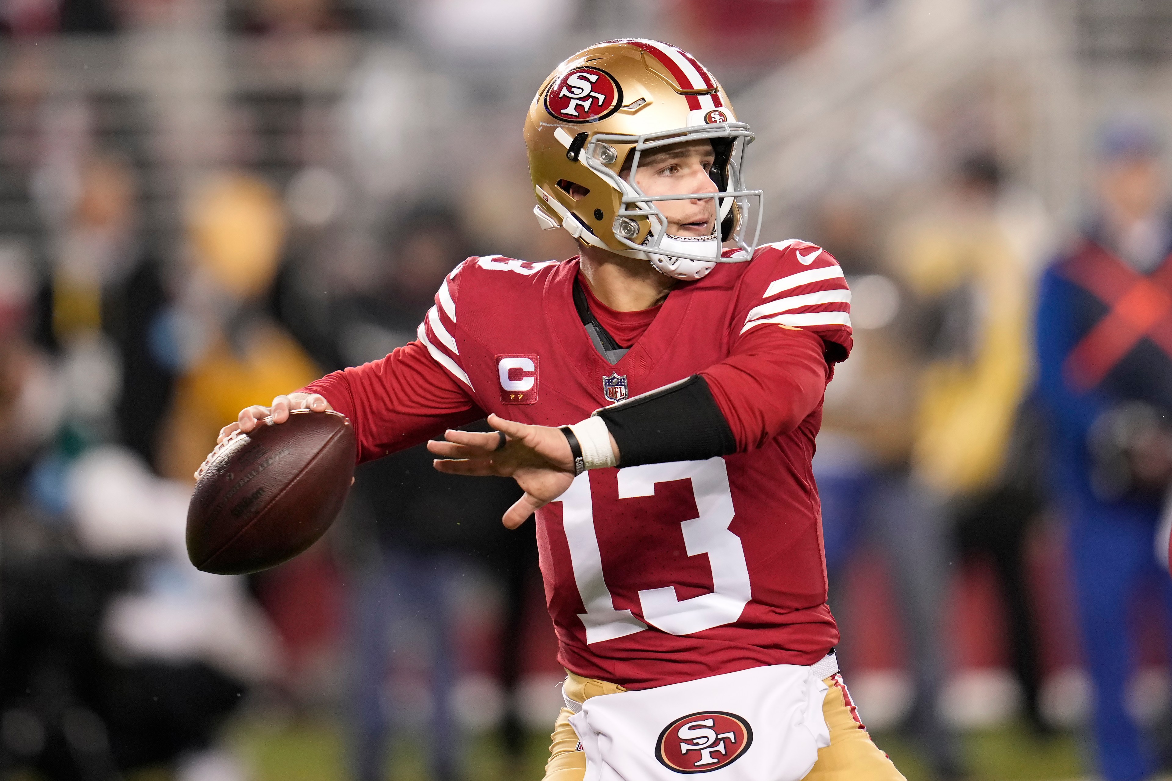 A football player in a red and gold uniform, number 13, is poised to throw the ball. He wears a helmet and a left arm sleeve, with a focused expression.