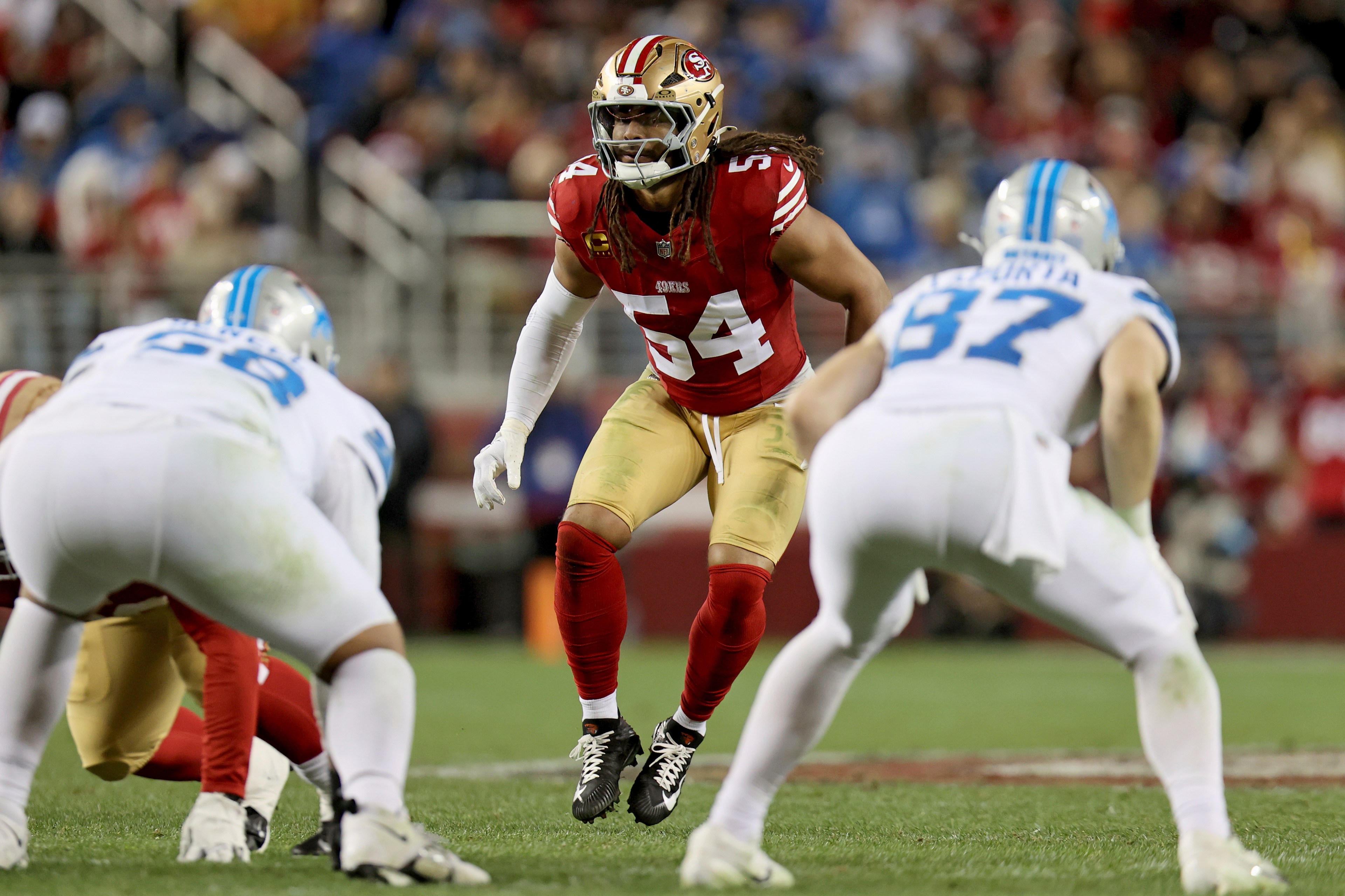 A football player in a red and gold jersey is poised and ready on a field, surrounded by players in white and blue uniforms, during a game.