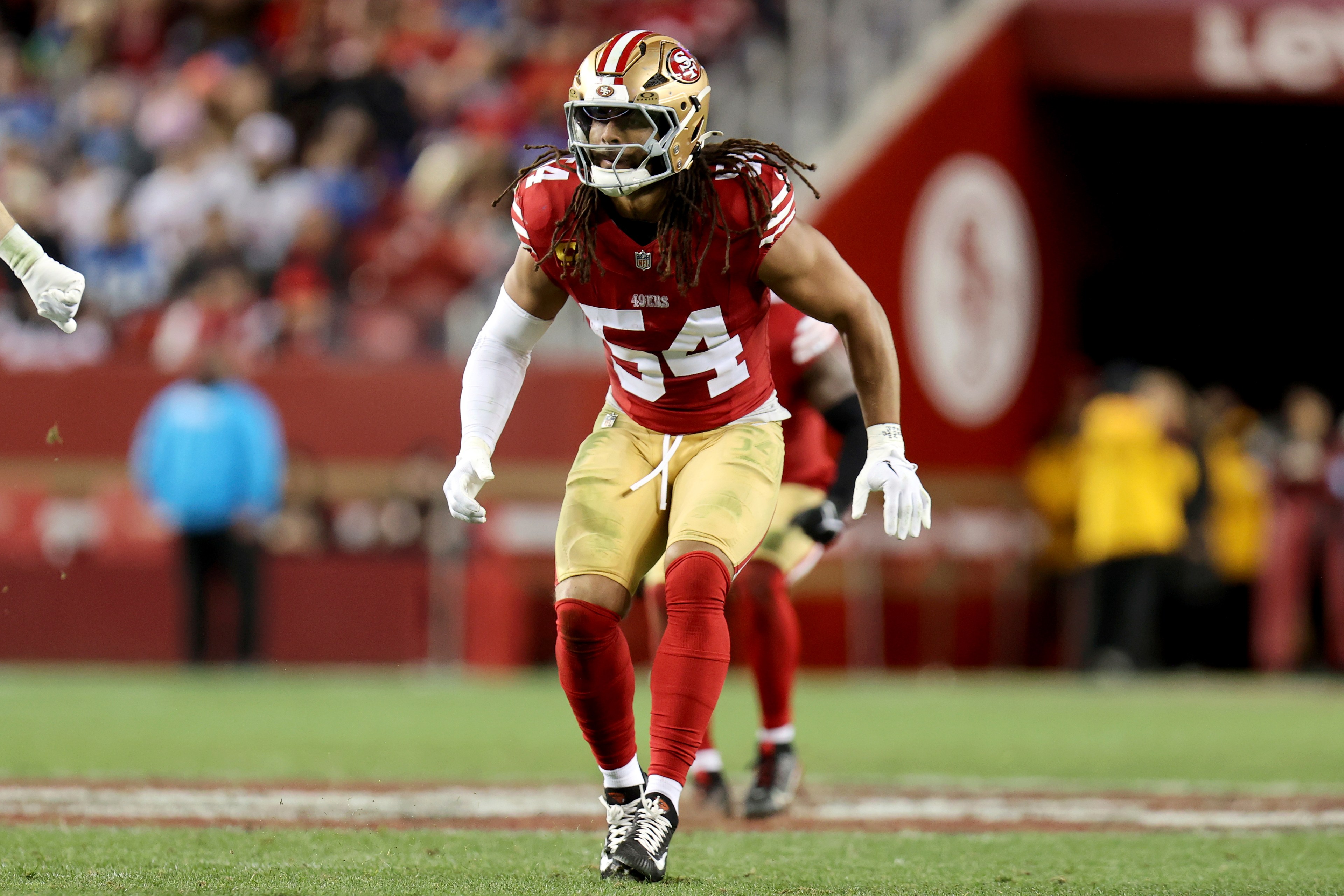 A football player in a red and gold uniform is mid-action on the field, wearing a helmet and visor. The background is a blurred stadium.