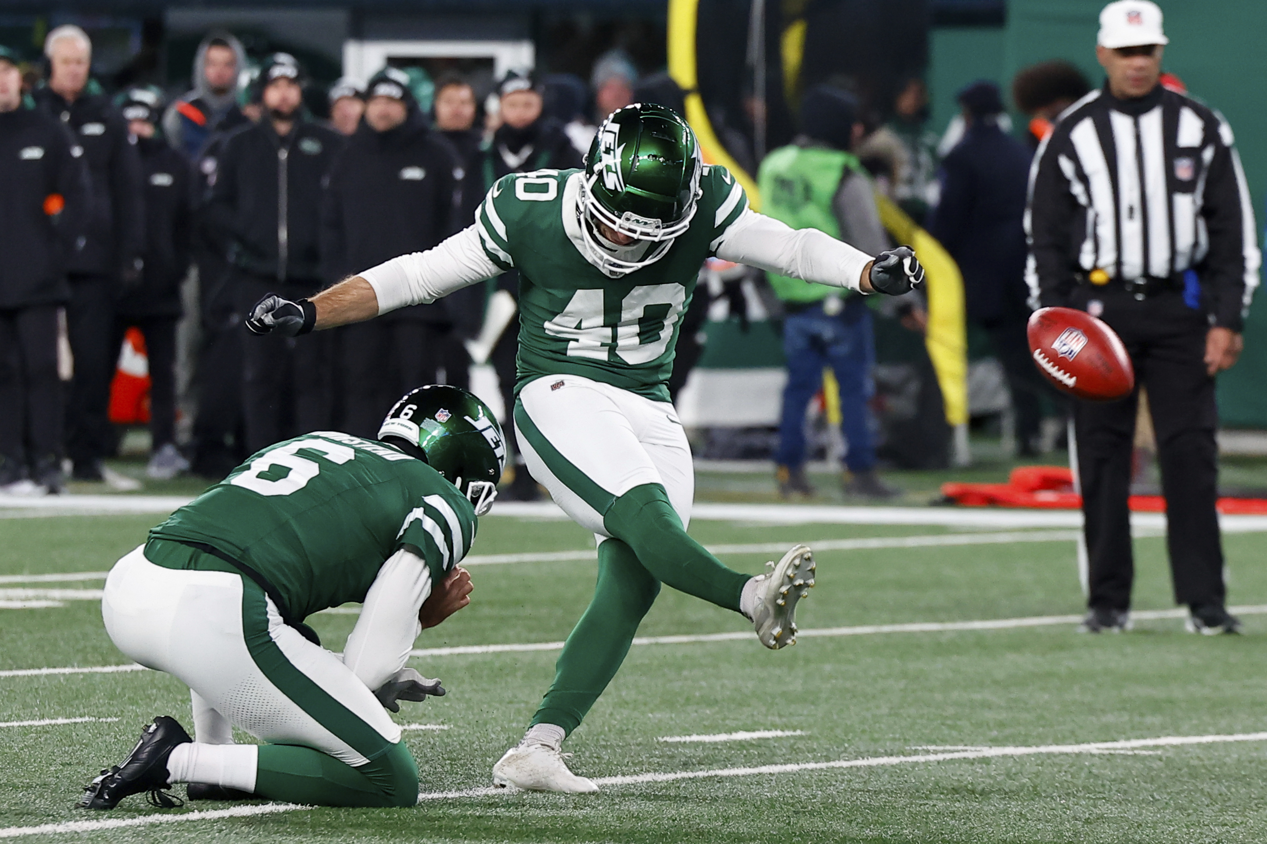 Two football players in green uniforms are on a field. One is holding the ball on the ground, while the other is kicking it. Spectators and referees are in the background.