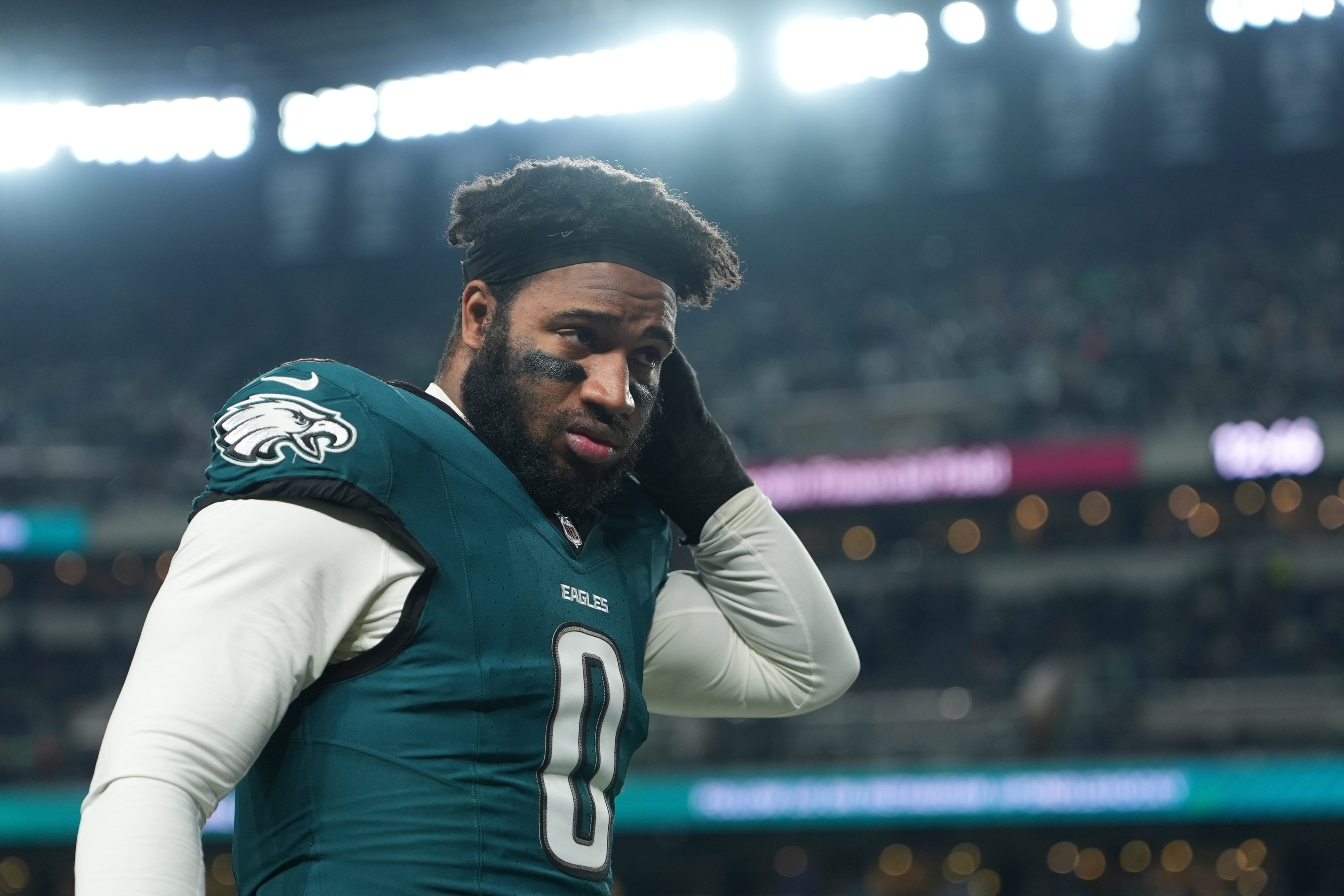 A football player in a green and white uniform with the number 0 adjusts his hair under stadium lights. He has black eye paint and looks focused.