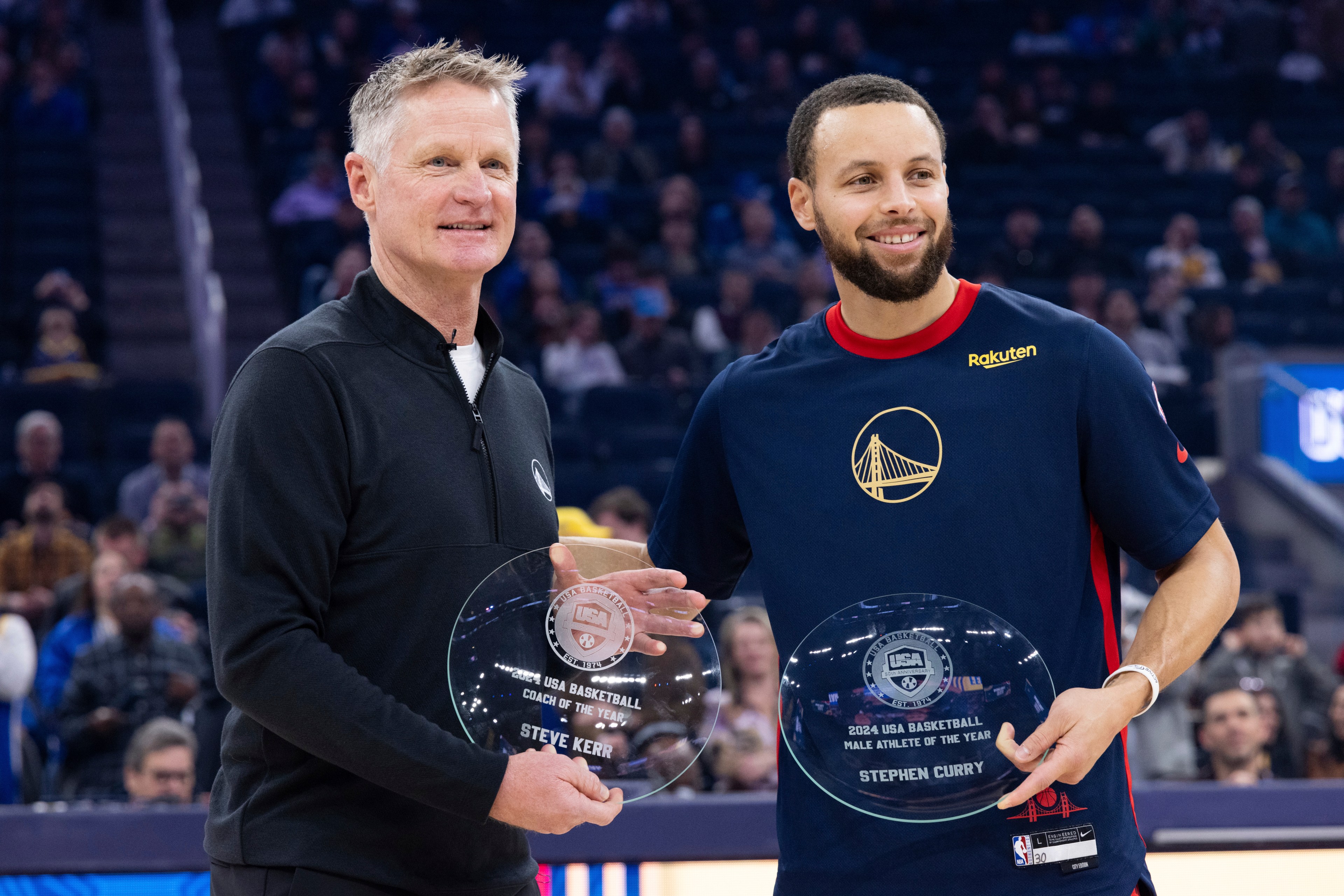Two men stand smiling, holding glass awards. One wears a black jacket, the other a navy shirt with a logo. They're in a sports venue with a crowd behind them.