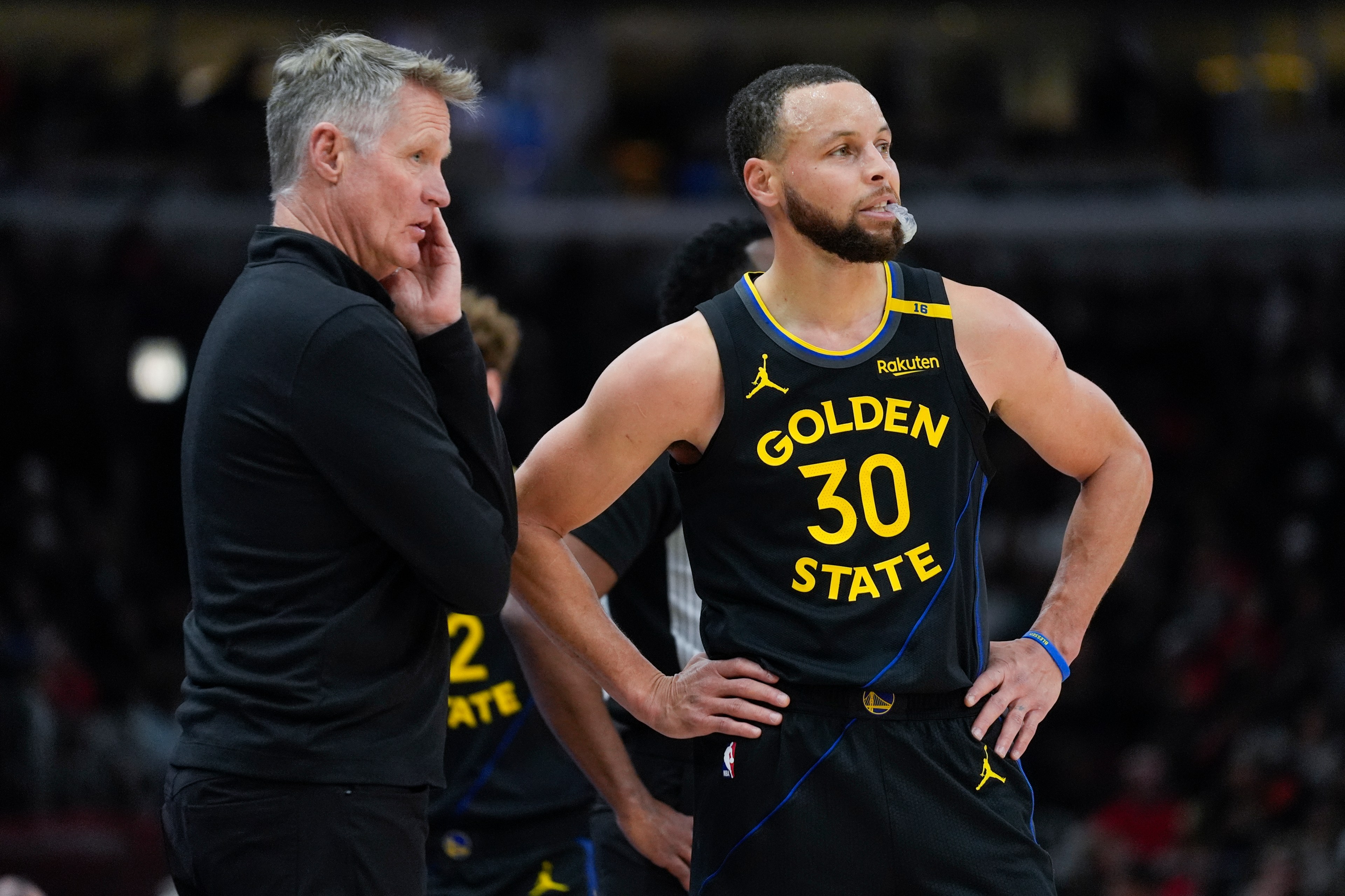 A basketball player in a "Golden State" jersey stands with hands on hips, alongside a man in a black jacket. The focus seems to be on a game or strategy.