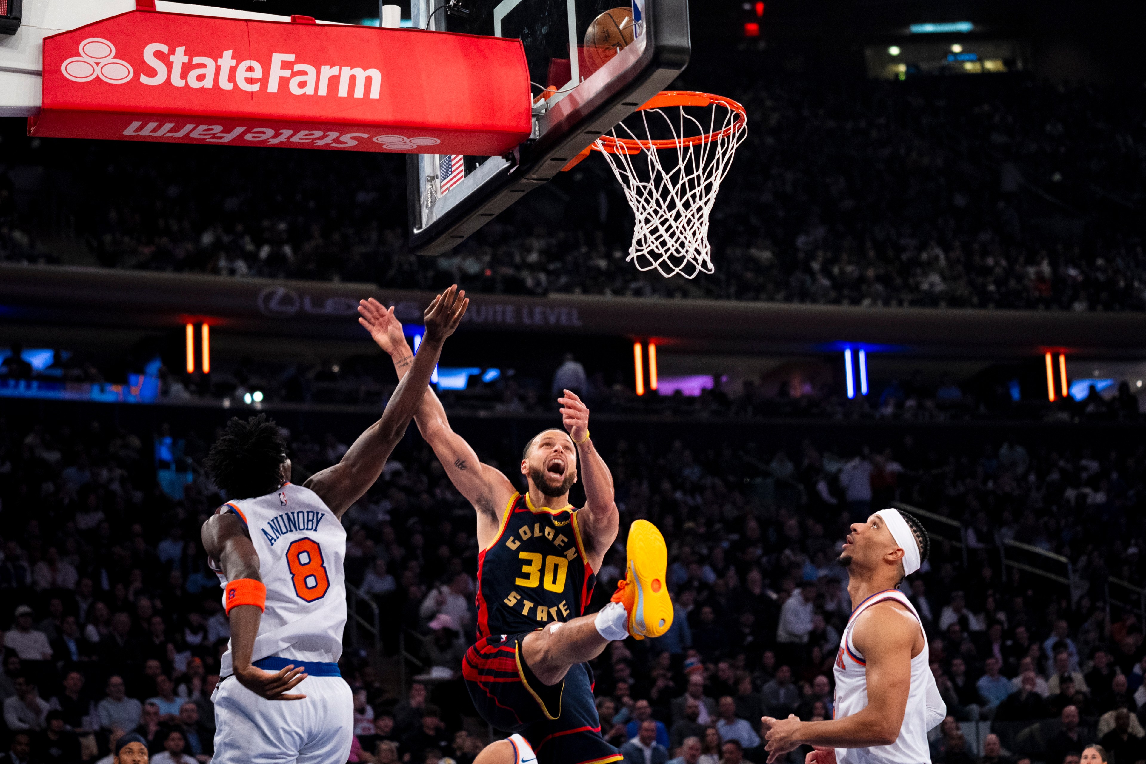 A basketball player in a dark jersey leaps towards the hoop, ball in hand, as opponents in white jerseys watch, under a bright red State Farm banner.