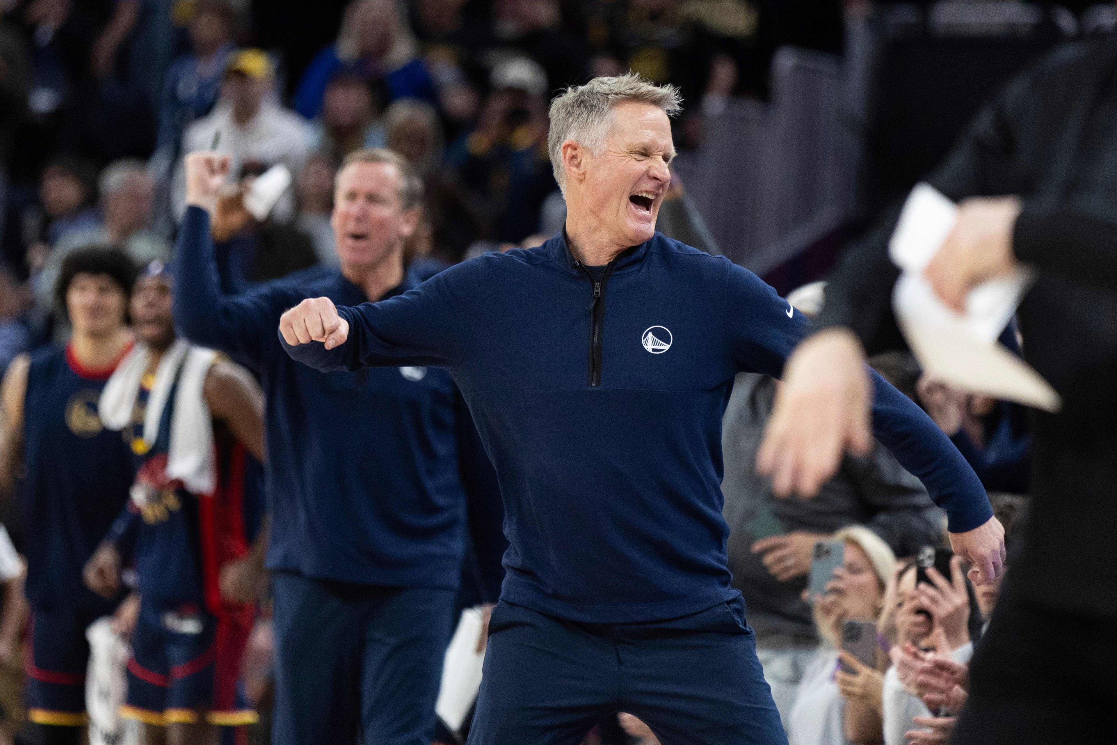 A man in a dark sports jacket is enthusiastically celebrating in a crowded basketball arena, surrounded by cheering fans and team members.